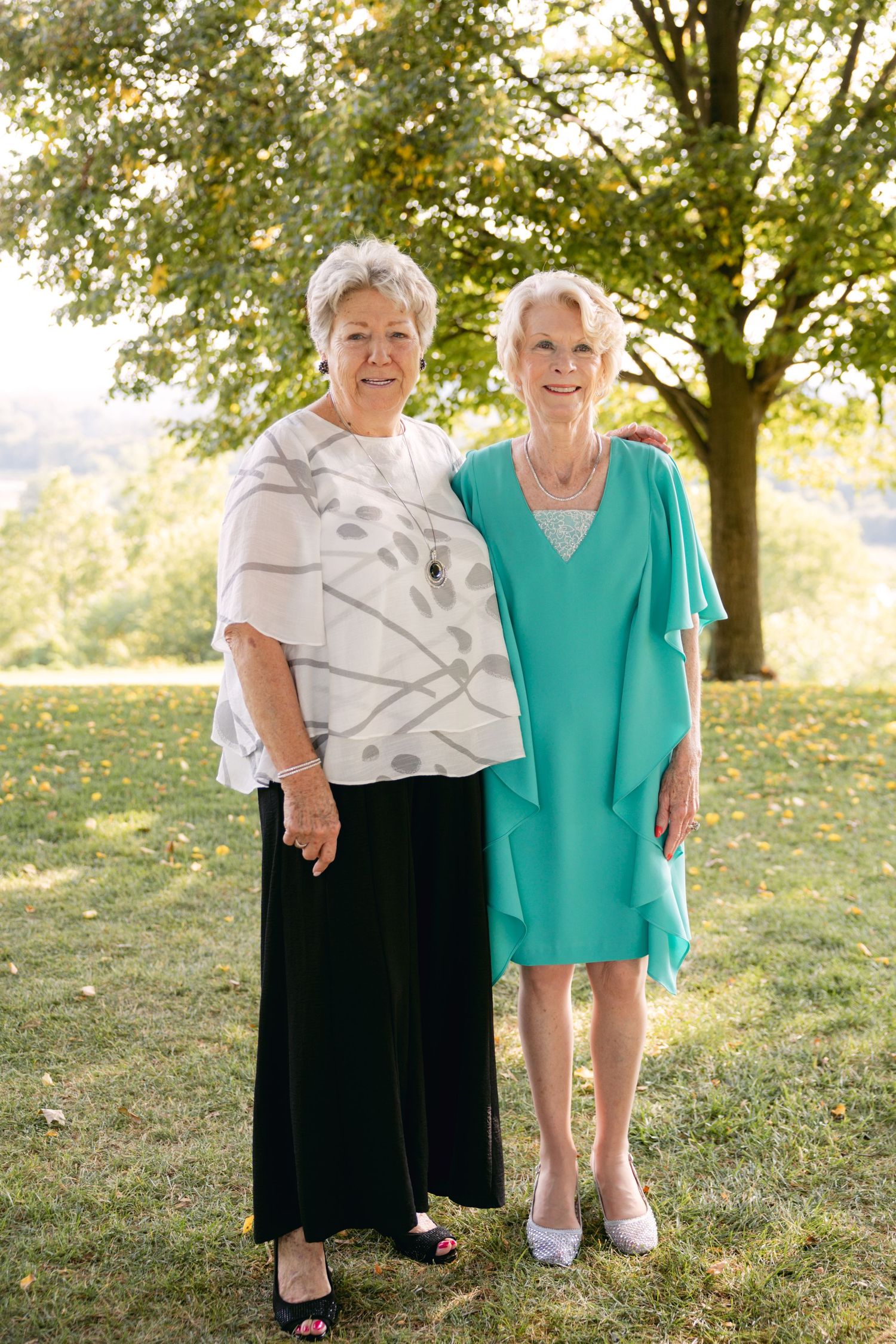 Two elegant women in casual formal wear pose together on sunny lawn.