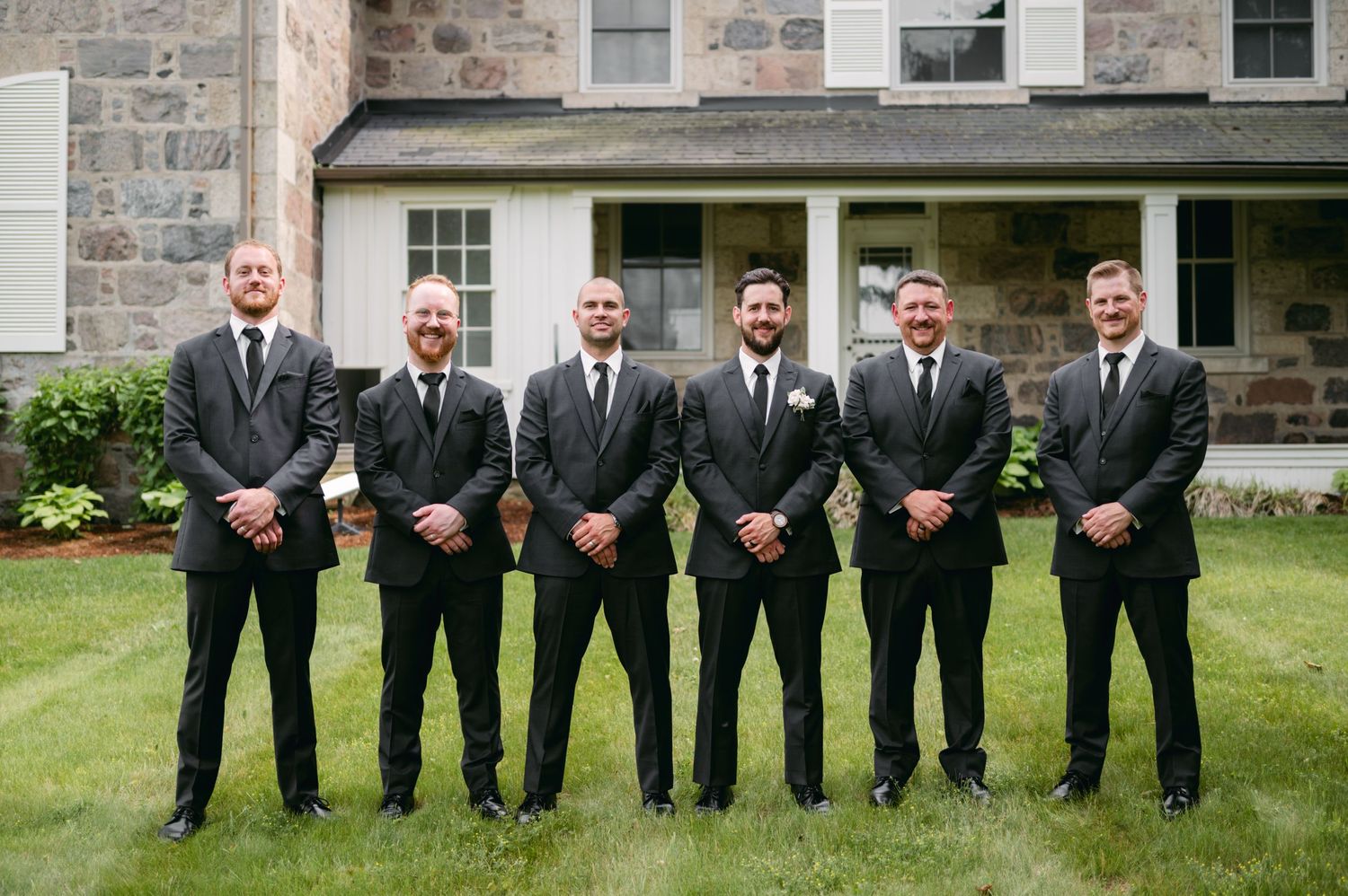 Groomsmen in black suits and ties stand in formation on green lawn by stone building.