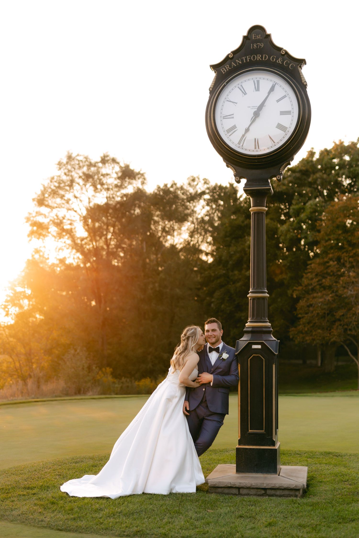 Newlyweds embrace beside ornate black clock post during sunset wedding photography session.