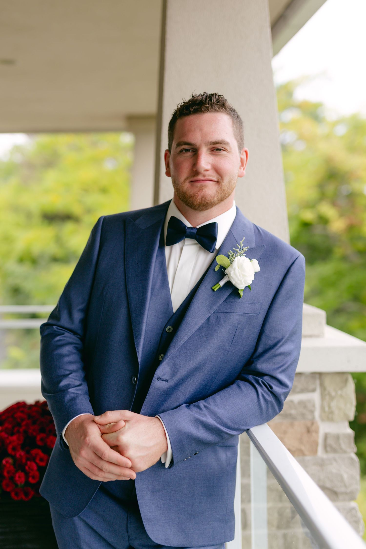 Person in navy suit with white boutonniere poses on covered porch with red flowers.
