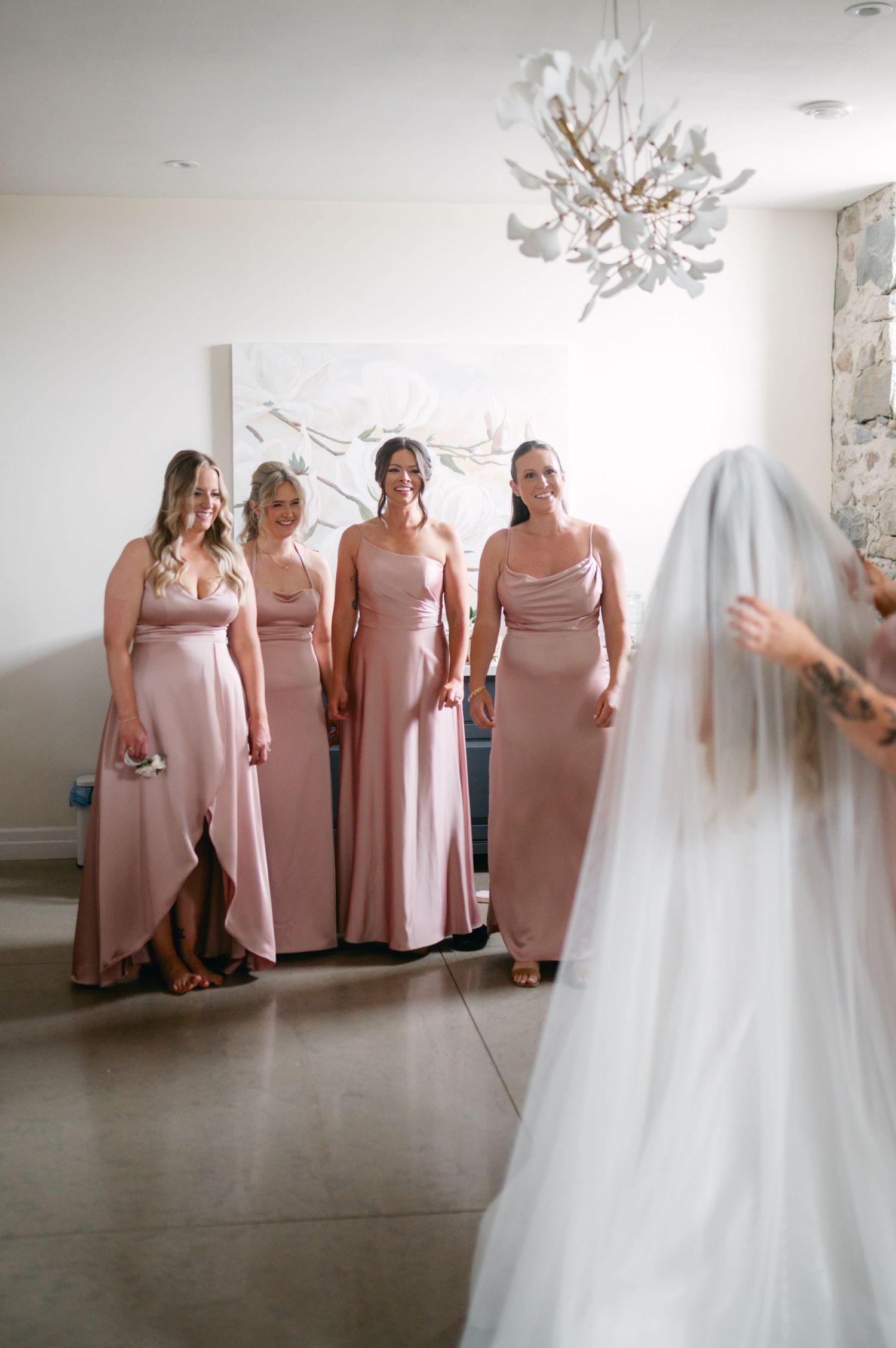 Bridesmaids in blush pink dresses smile at the bride in her white wedding gown and veil.