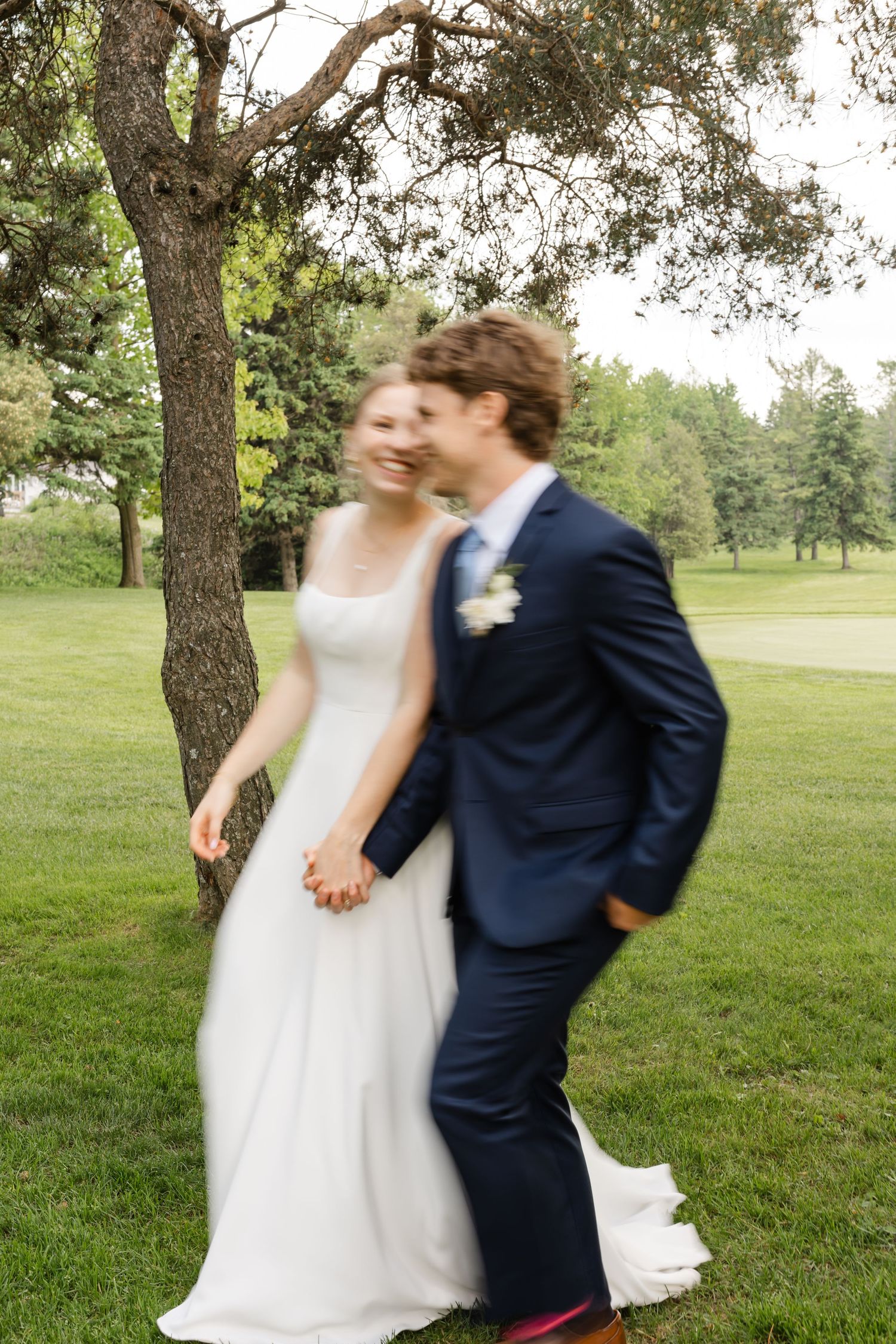 A couple in wedding attire spins and dances playfully on a lush green lawn.