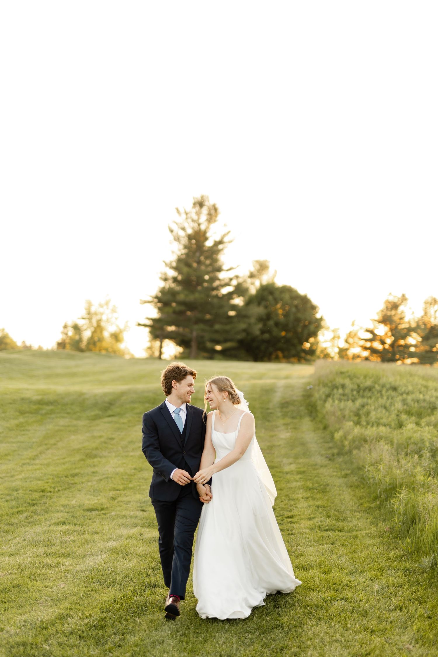 Wedding couple walks playfully through a grassy hillside at sunset.