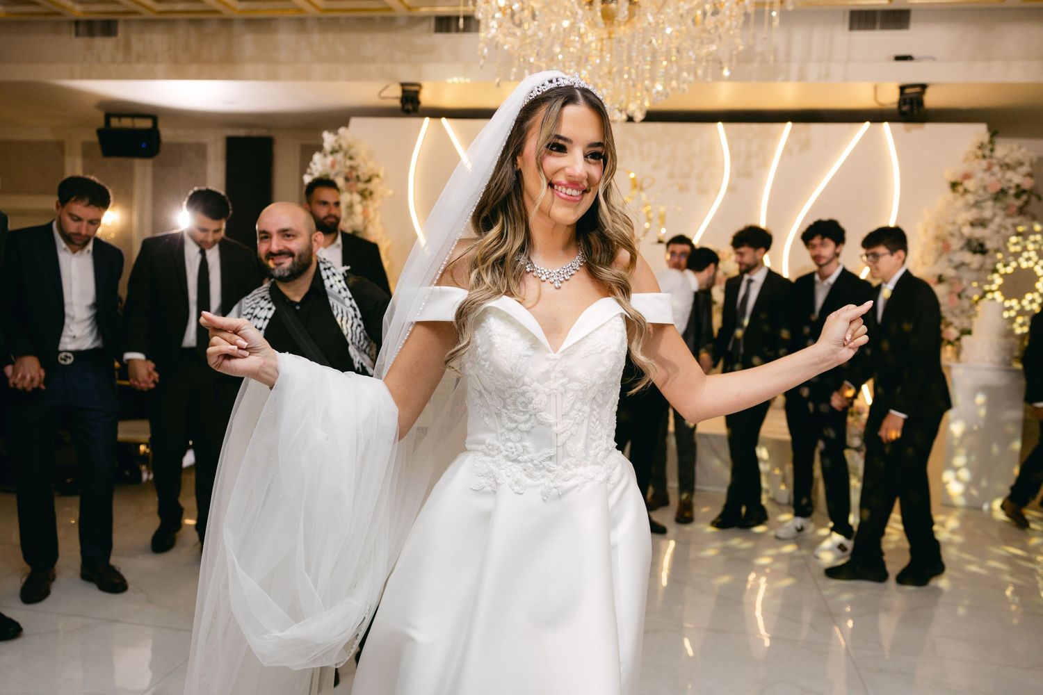 Wedding celebration in a modern white ballroom with guests dancing under crystal chandeliers.