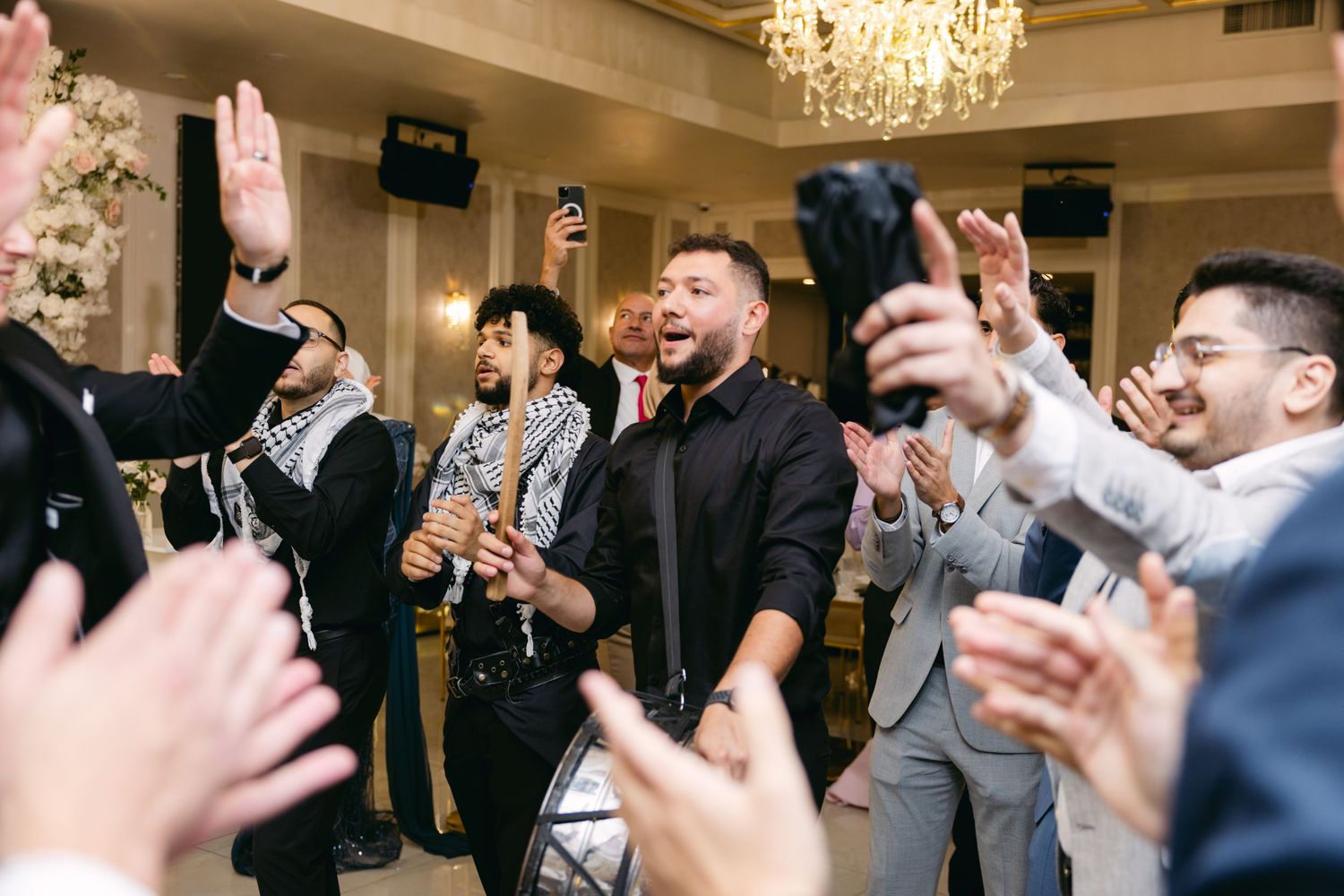 Wedding guests raise their hands in celebration during a festive reception under crystal chandeliers.