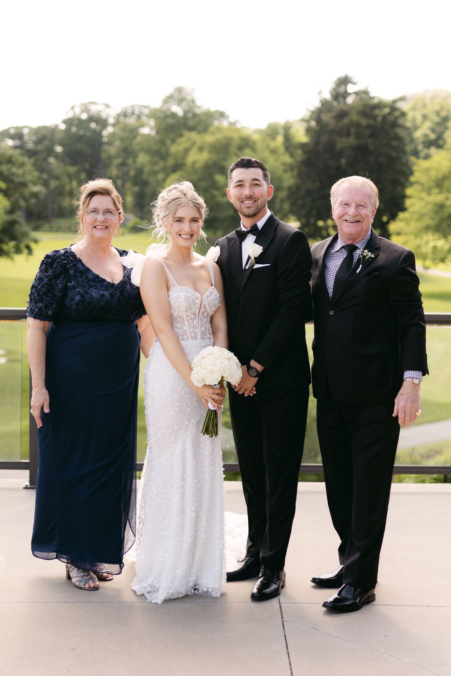 Wedding party members pose formally in black attire with bride in white lace dress on outdoor patio.