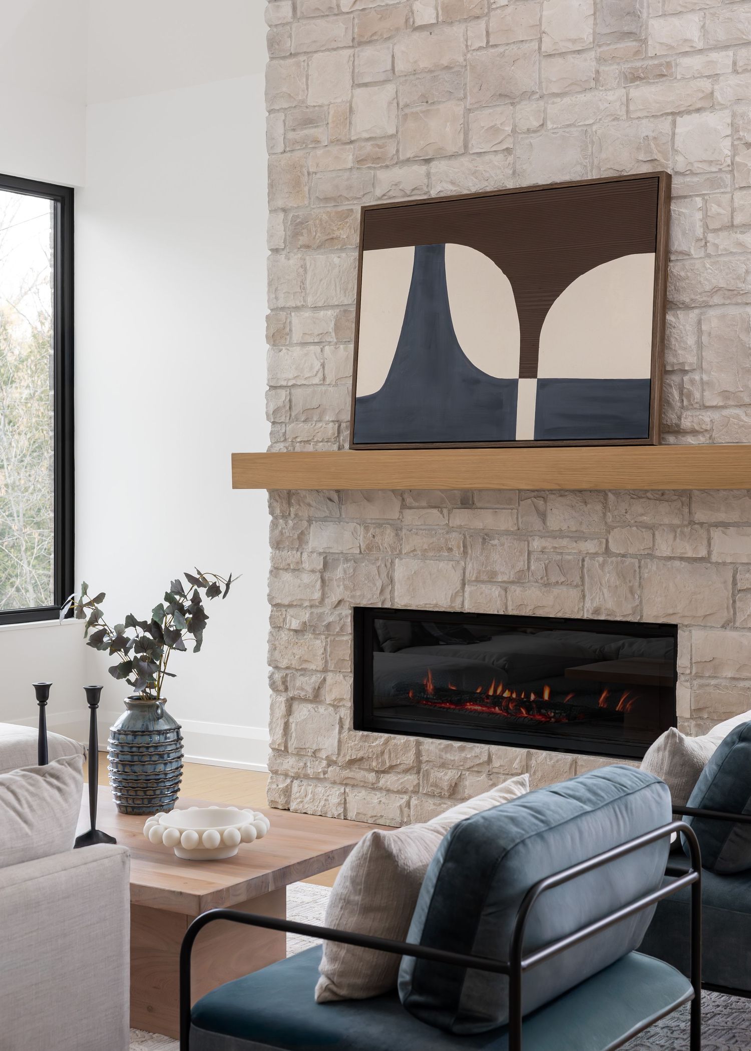 Modern living room features a stone fireplace with abstract art, blue accent chair and natural decor elements.