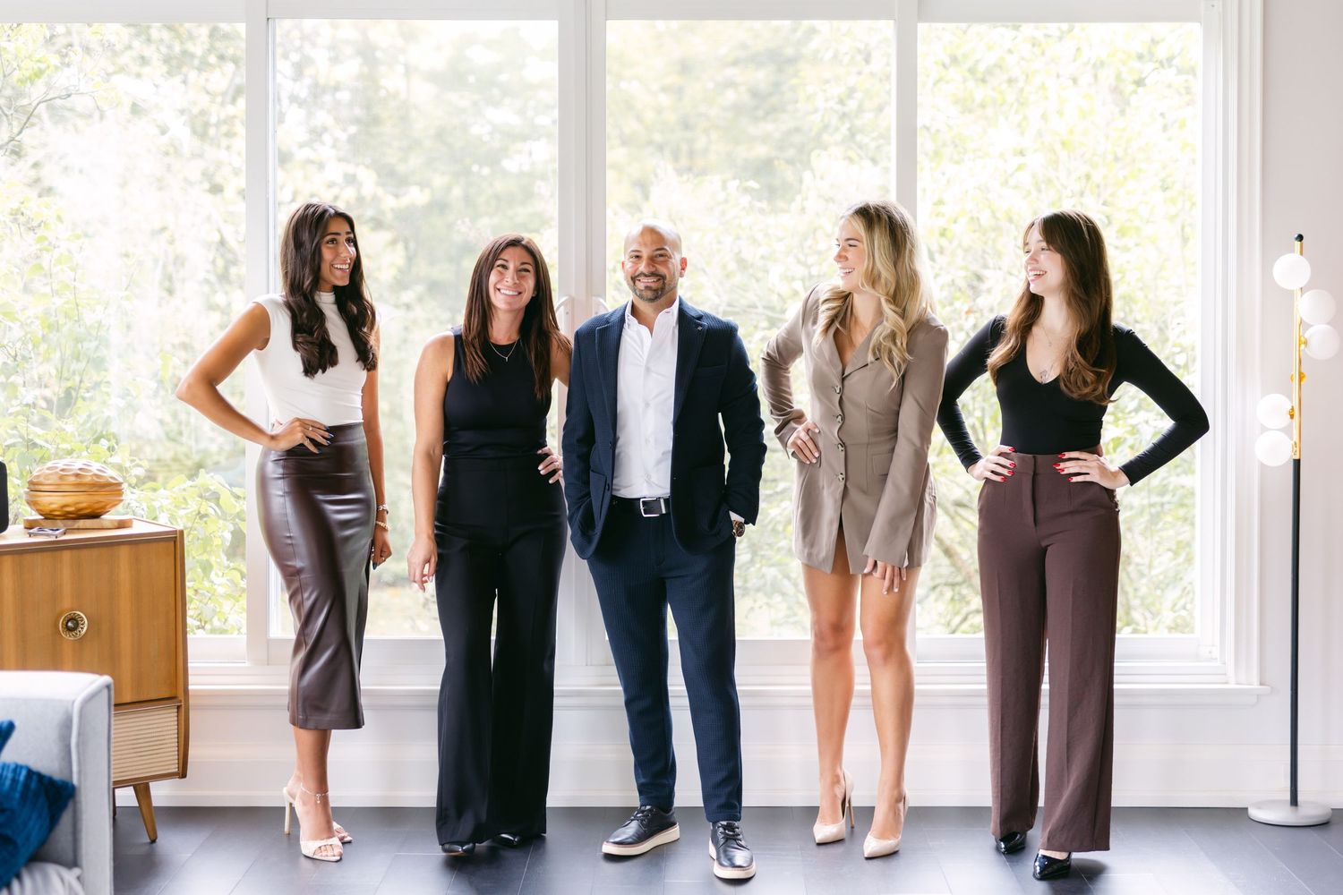 Professional business team members stand together in a row wearing formal attire in a bright modern office setting.