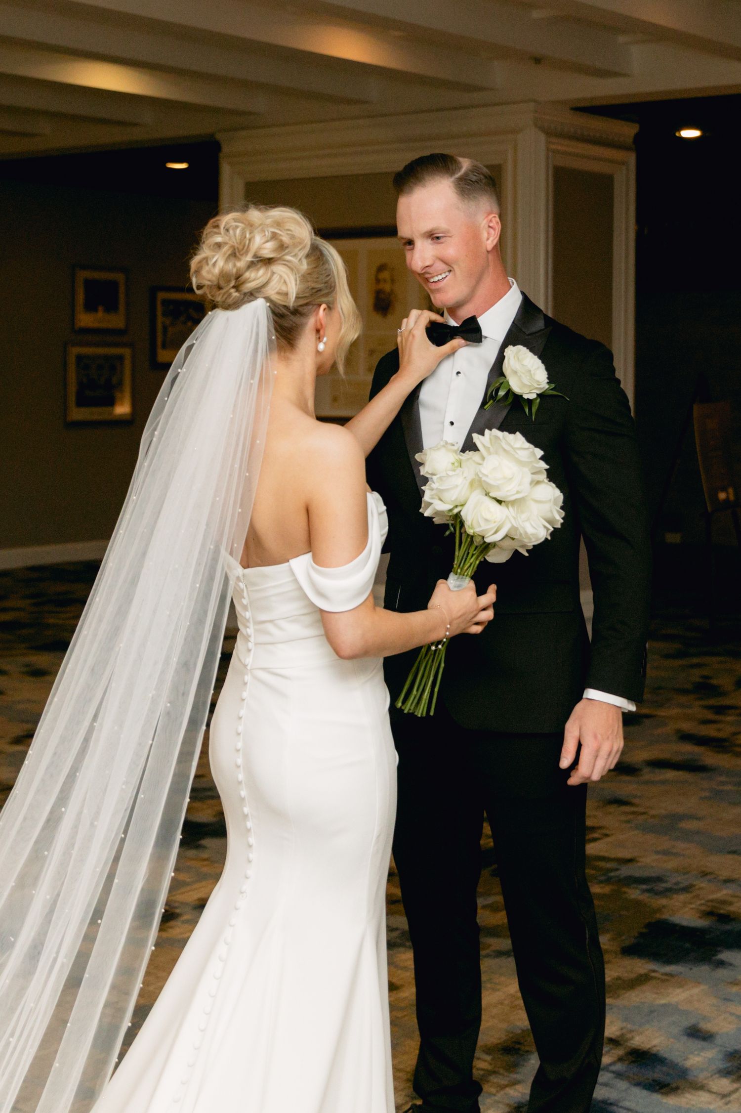 Bride in off-shoulder white gown adjusts groom's bow tie as he smiles, holding white rose bouquet indoors.