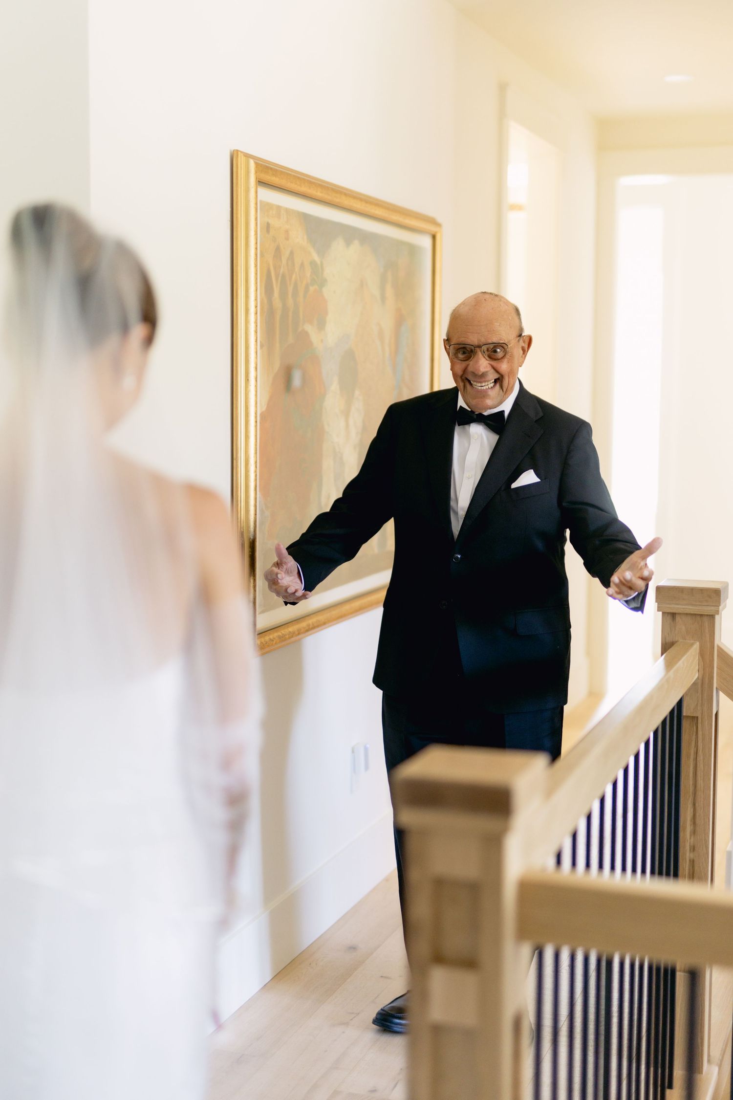 Father in black tuxedo reacts with joy and open arms seeing bride in white dress during wedding first look.