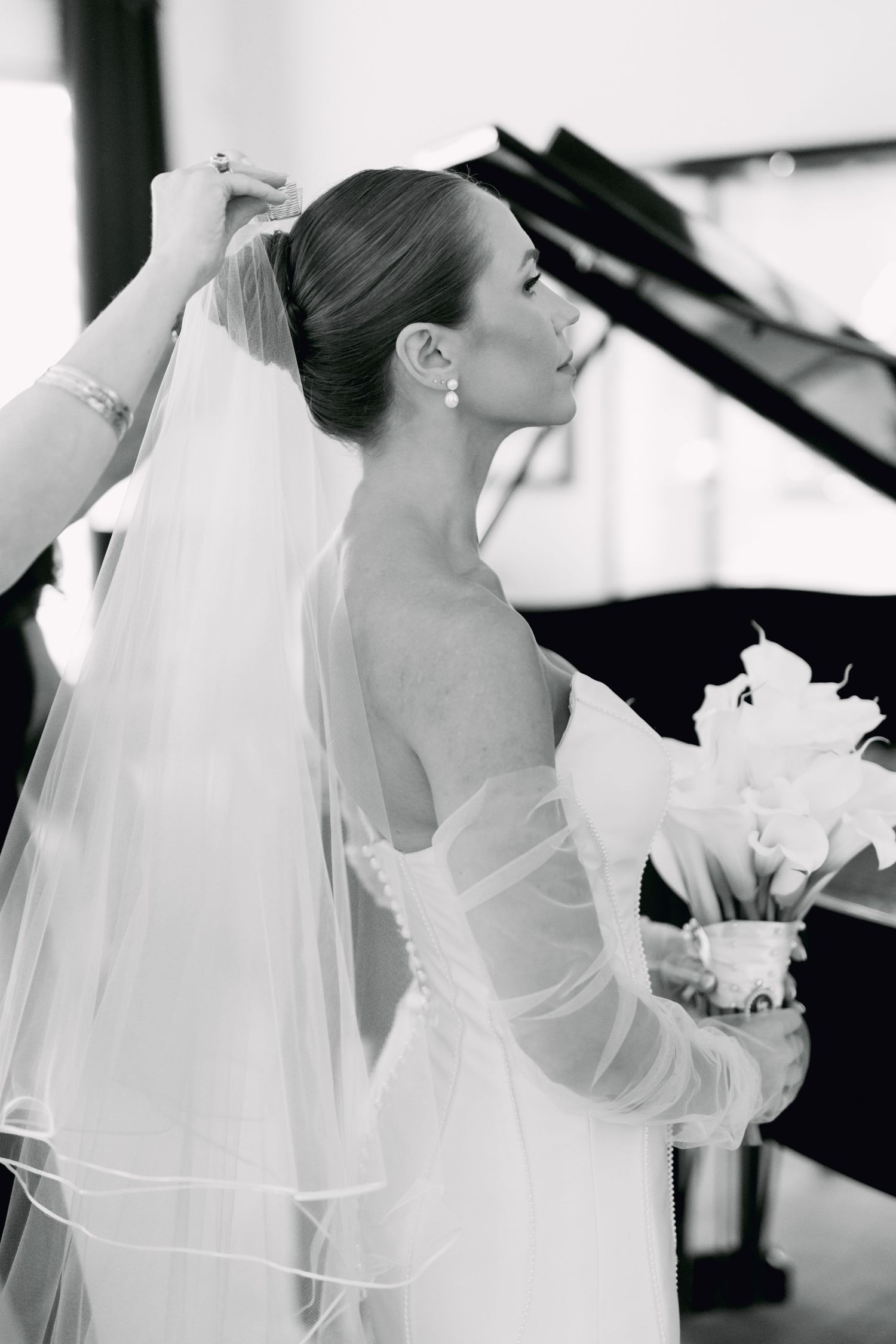 Black and white photo of bride in strapless gown holding bouquet while veil is adjusted near grand piano.