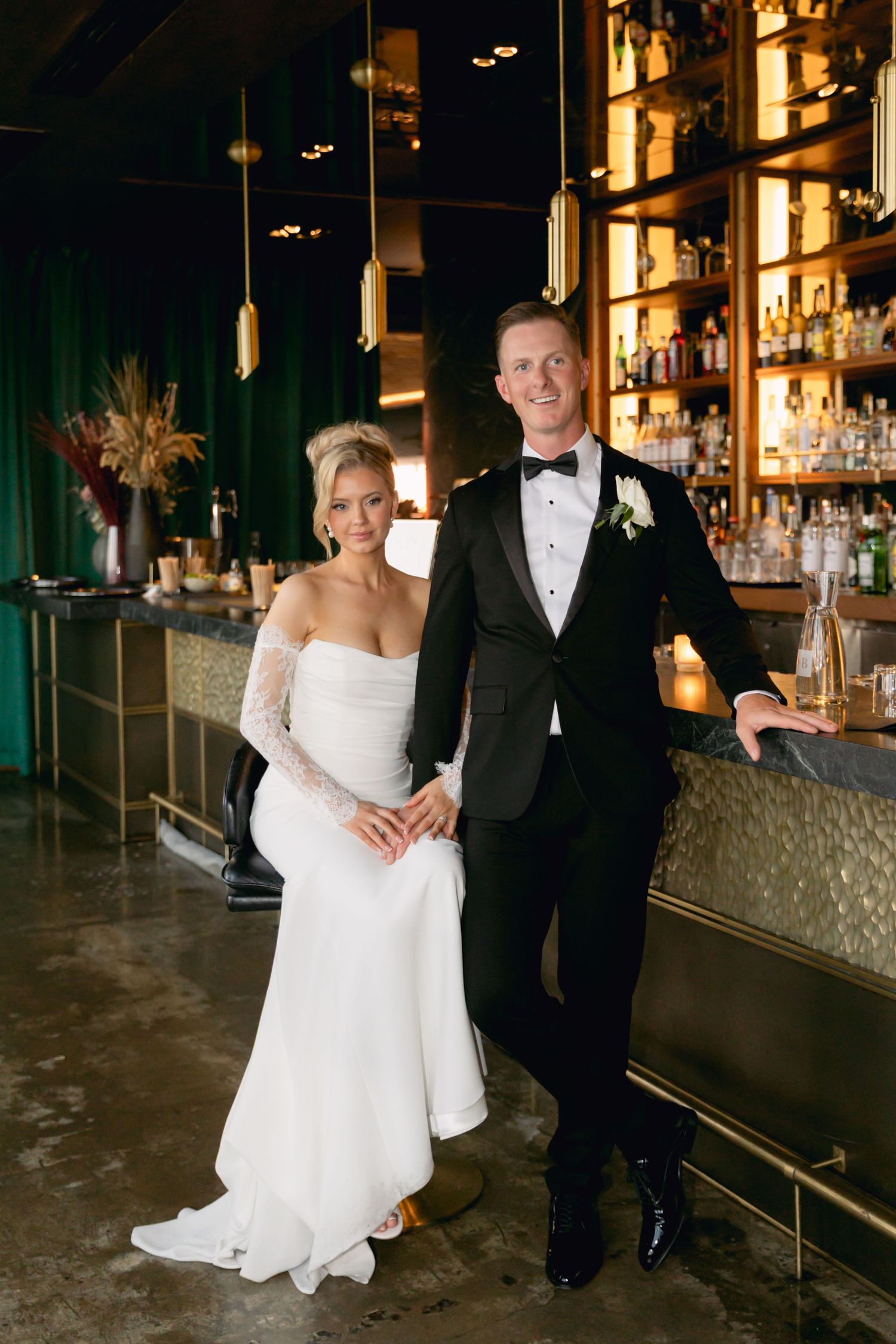 Bride and groom posing together at stylish bar with green velvet backdrop and gold pendant lights.