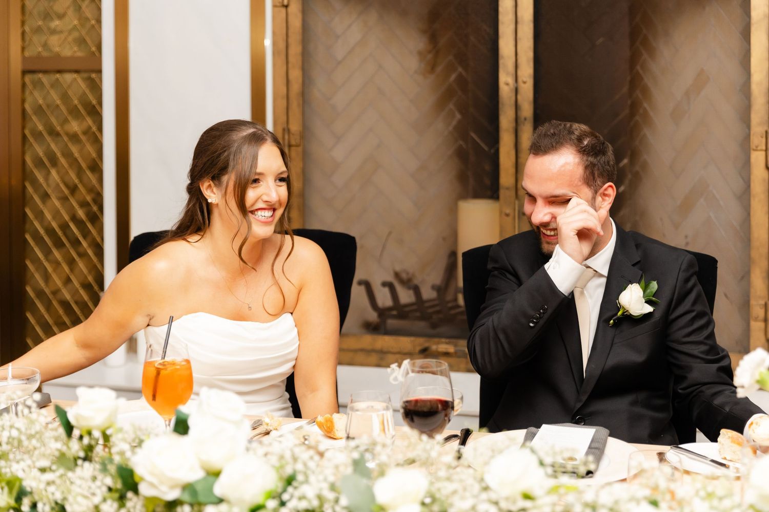 Bride laughs while groom wipes tears during heartfelt wedding reception speeches at head table.