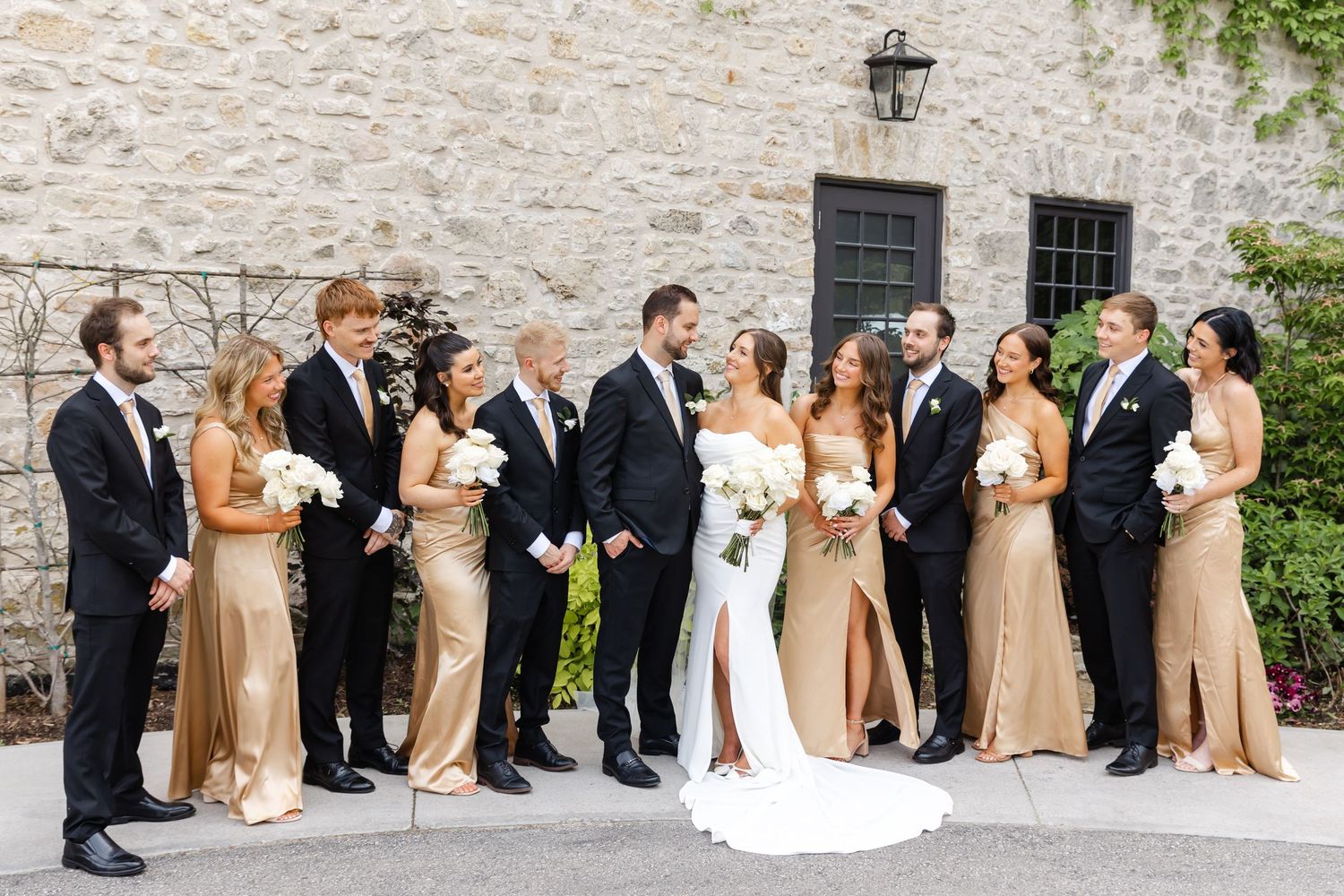 Wedding party in black tuxedos and gold bridesmaid dresses with bride in white gown holding bouquets outside stone building.