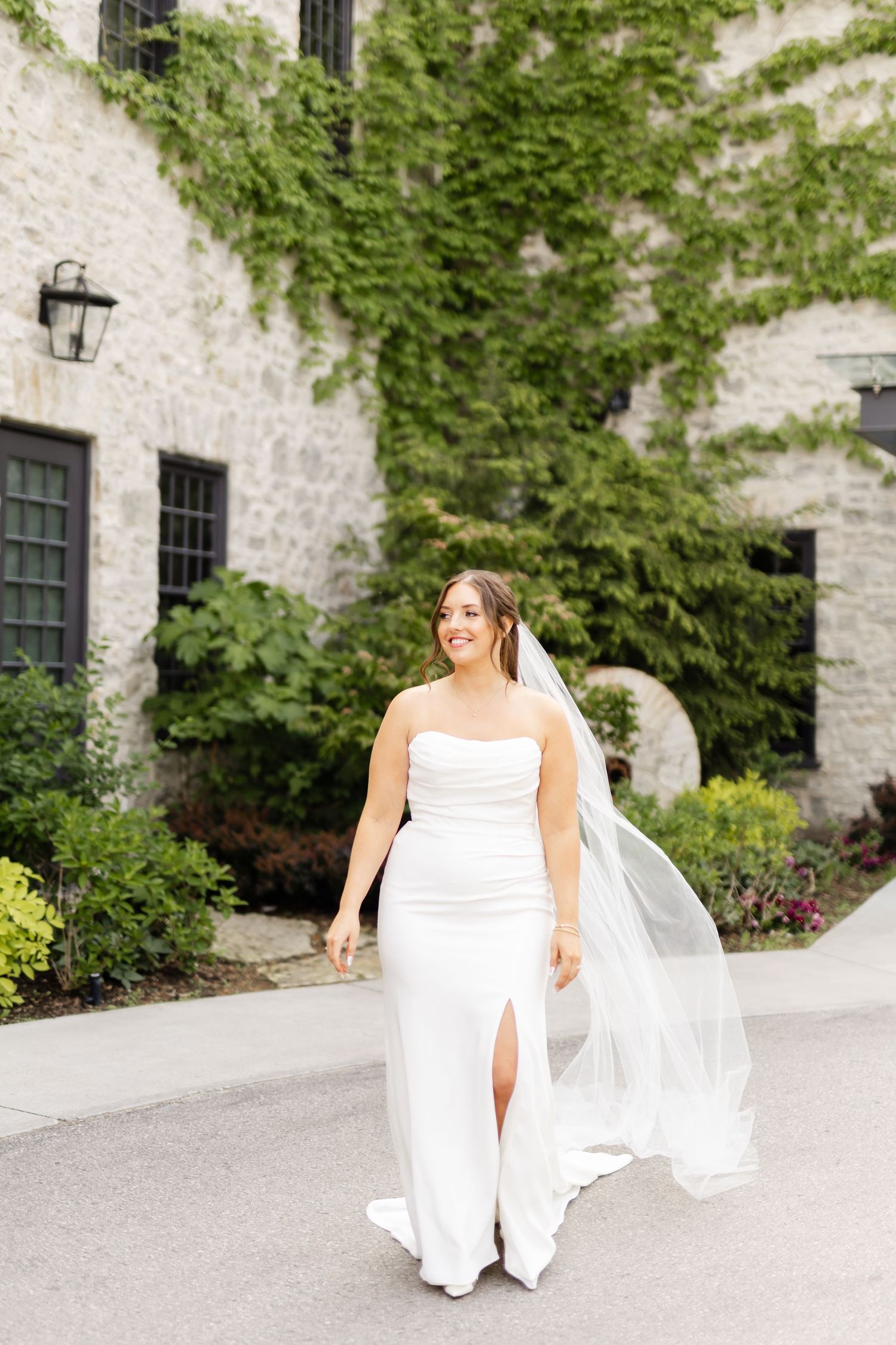 Bride in strapless white gown with flowing veil walks outdoors in front of ivy-covered stone building.