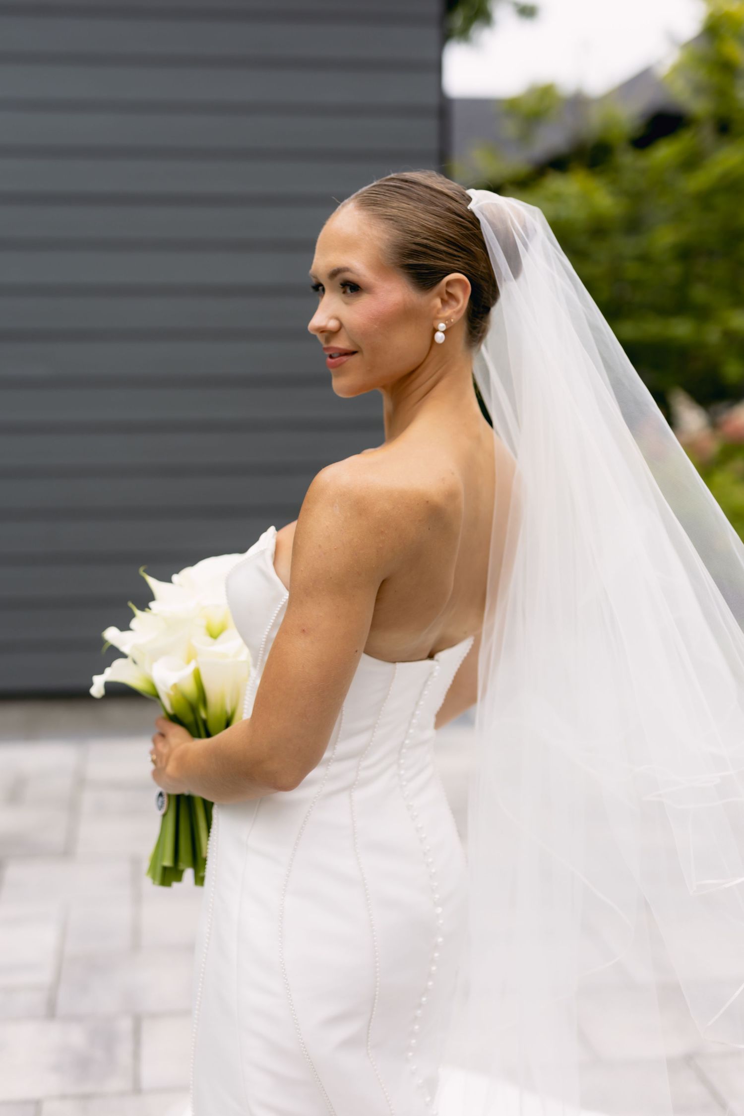 Bride in strapless white mermaid dress with veil holds white calla lily bouquet outdoors.