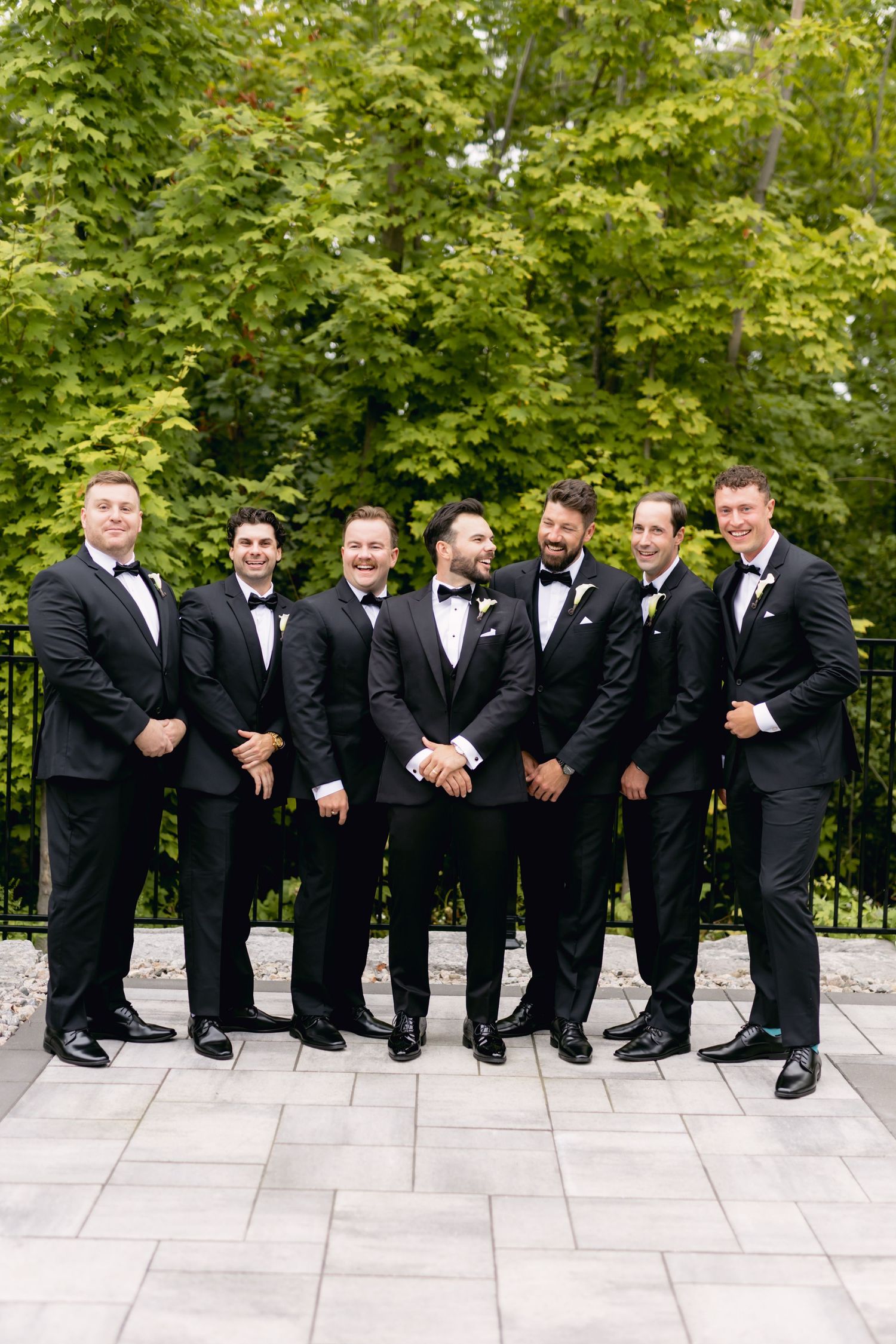 Groom and six groomsmen in black tuxedos posing outdoors on stone patio.