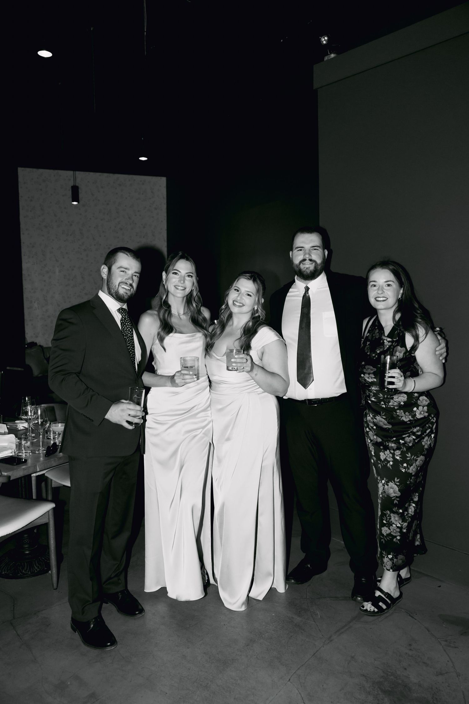 Black and white photo of five wedding guests smiling together holding drinks at a reception venue.