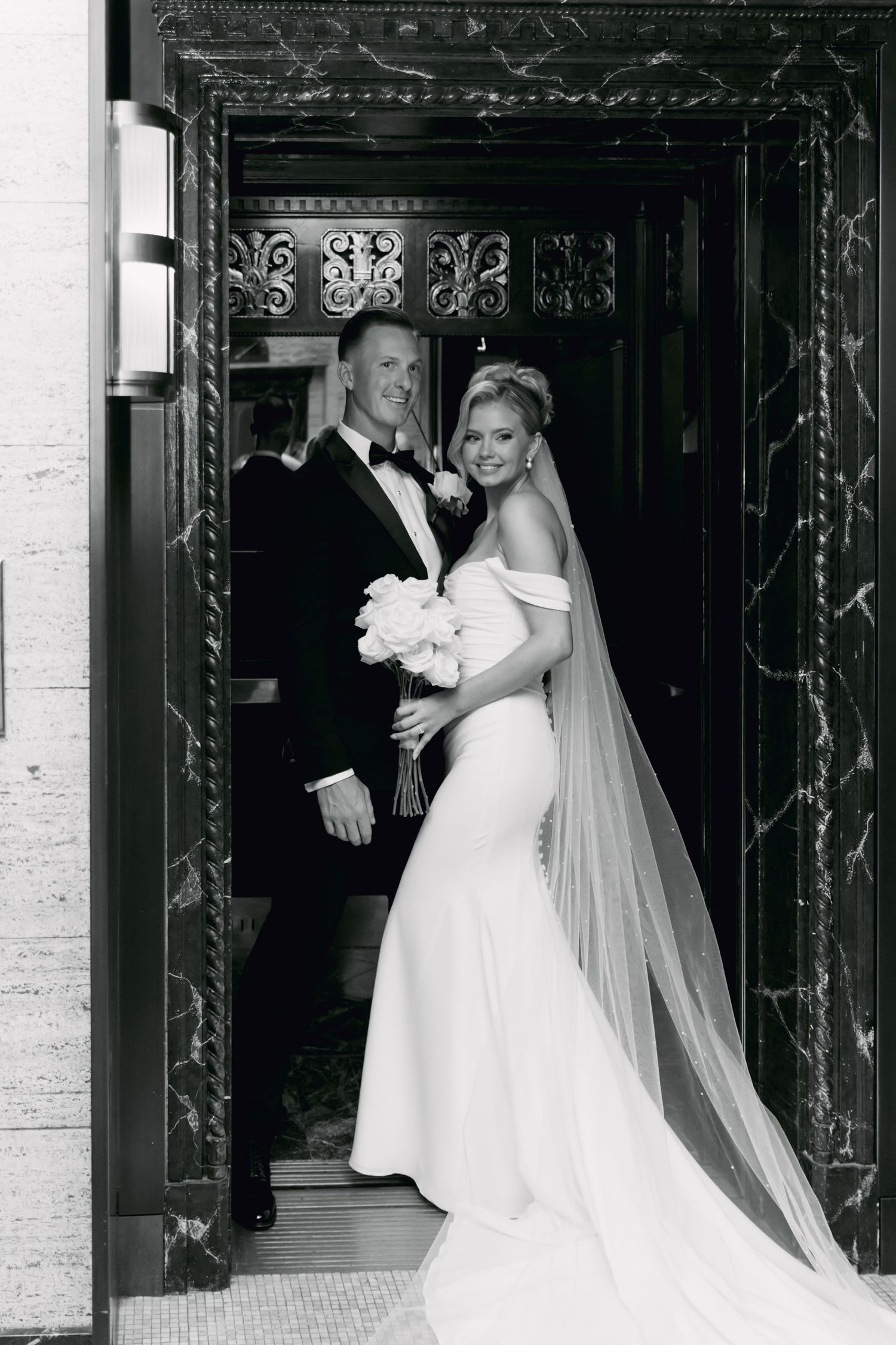 Black and white photo of bride and groom standing in an ornate doorway, bride holding white bouquet in fitted gown.