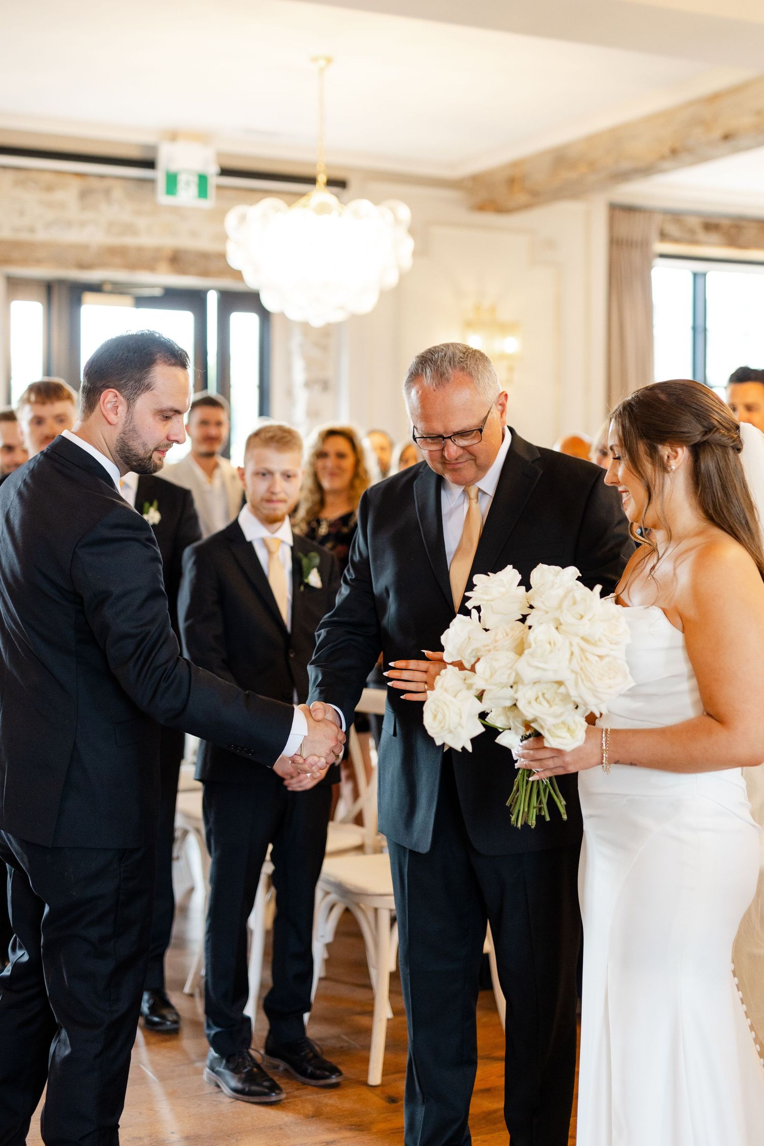 Groom shakes hand with father of bride during wedding ceremony while bride holds white rose bouquet.