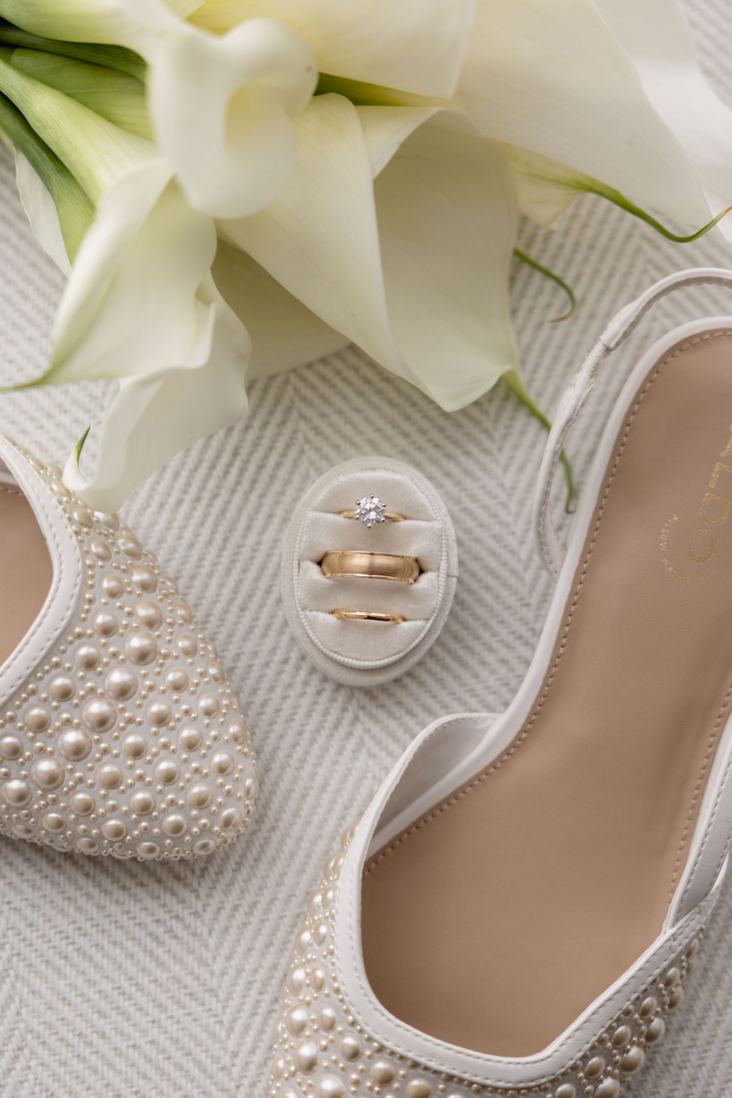 Pearl-adorned bridal heels beside gold wedding rings in a velvet box with white calla lily bouquet.