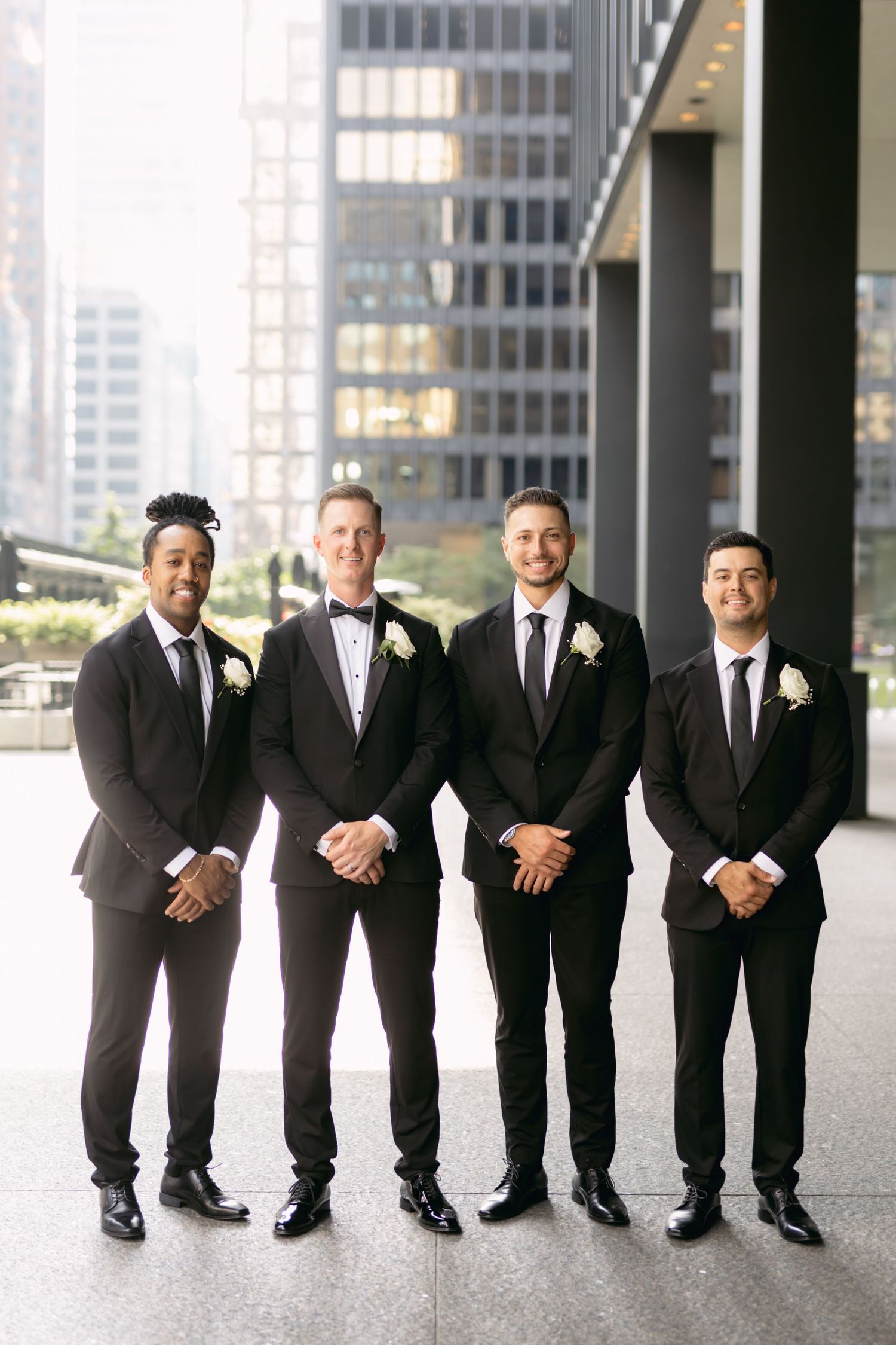 Four groomsmen in black suits with white boutonnieres pose together outside modern city buildings.