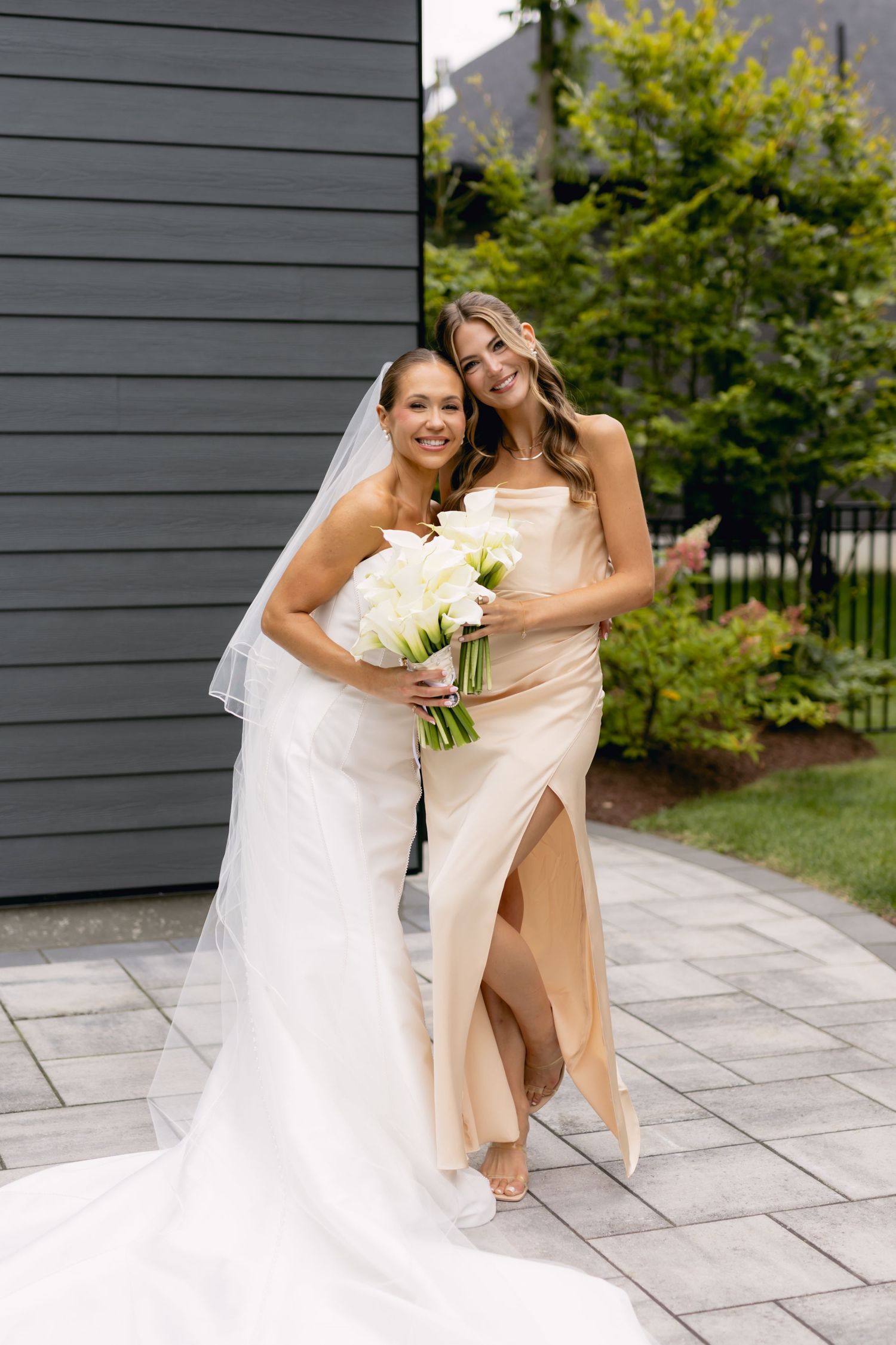 Bride in strapless white gown and veil poses with bridesmaid in champagne slit dress holding white calla lilies.