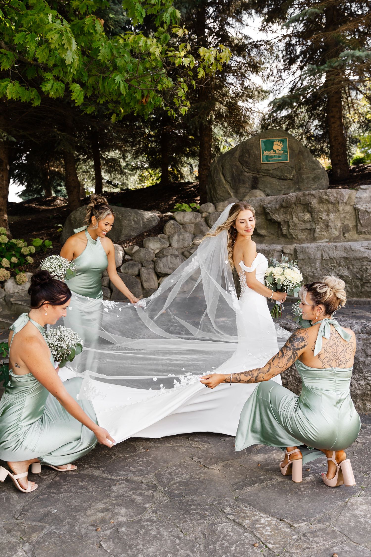 Bridal party in sage green dresses arranging the bride's veil on stone steps.