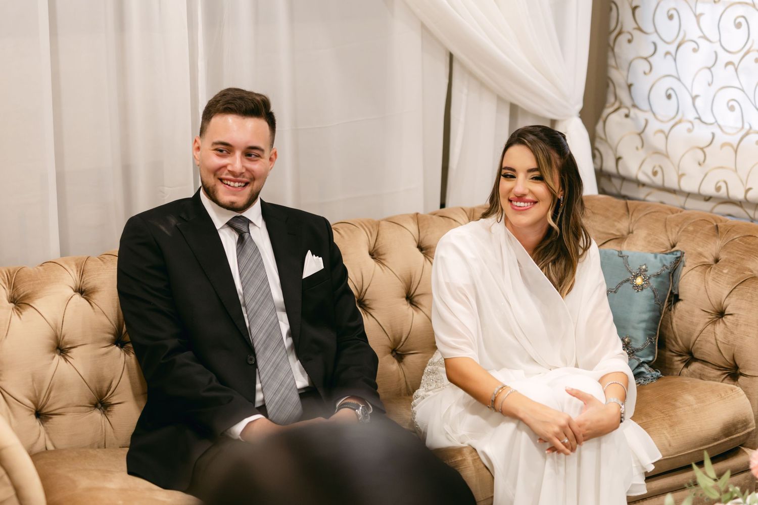 A couple sits together on a tan tufted couch during their wedding ceremony.