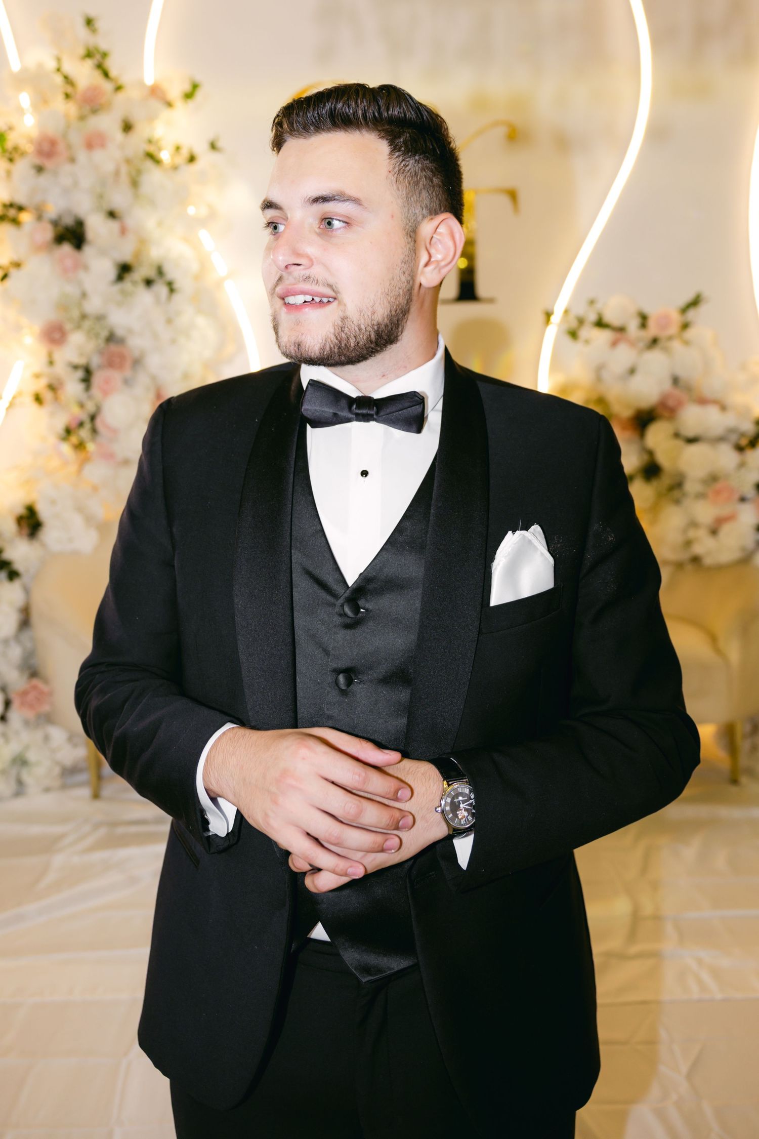 Groom in classic black tuxedo and bowtie poses in an elegant white floral setting.