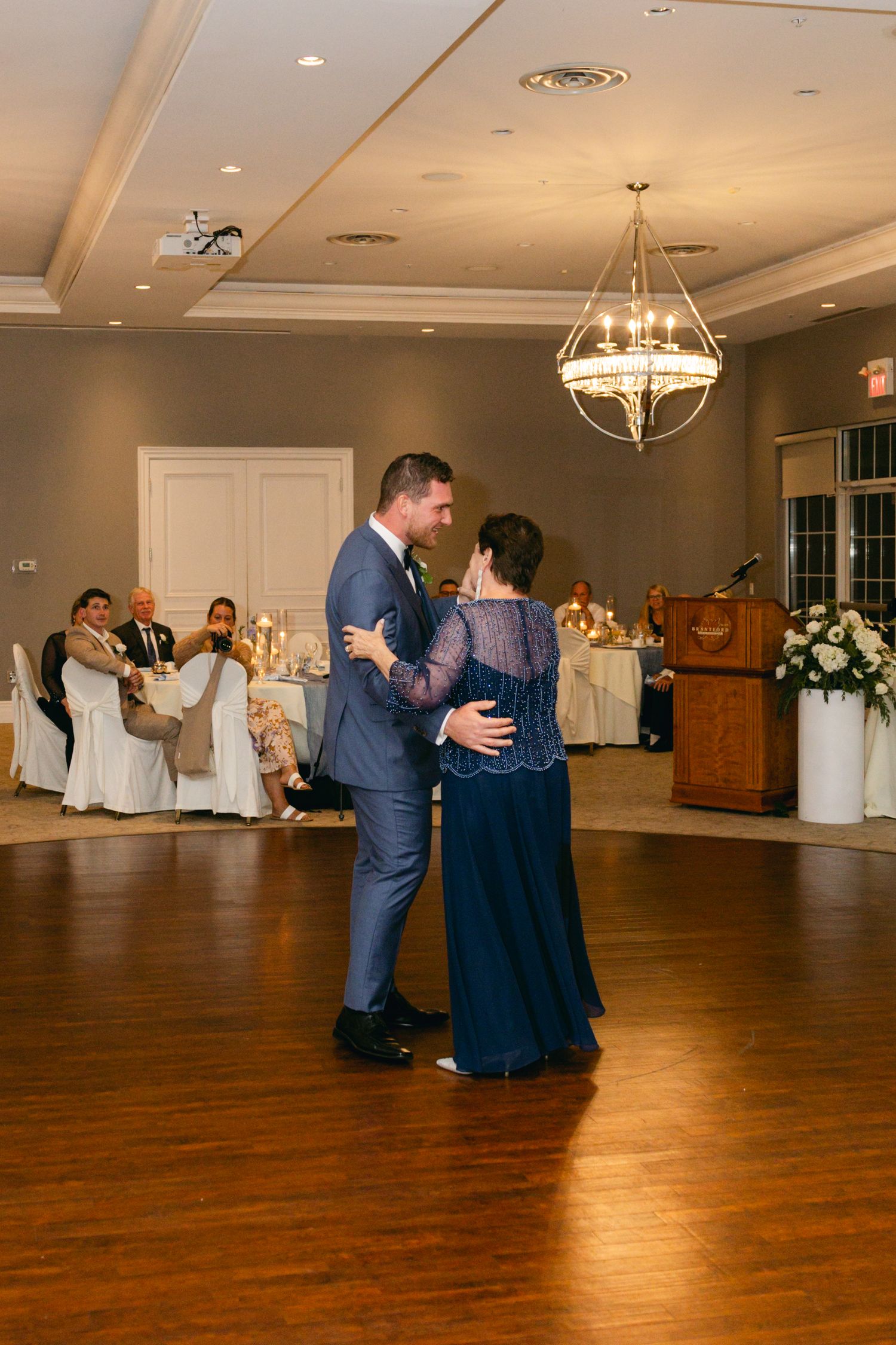 Wedding guests dancing together on a polished wooden dance floor under elegant chandeliers.
