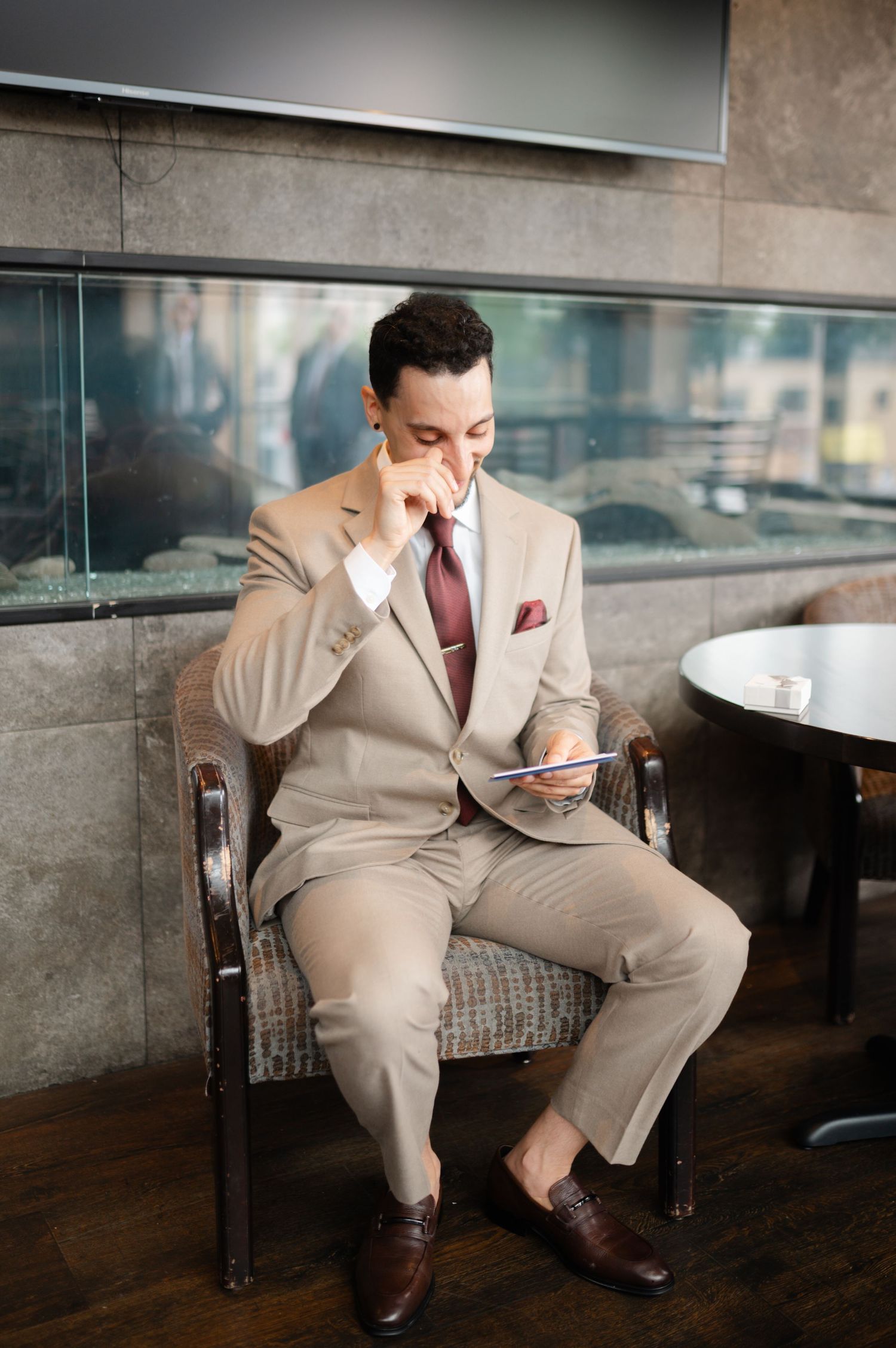 Professional in beige suit sits in armchair checking phone with thoughtful expression.