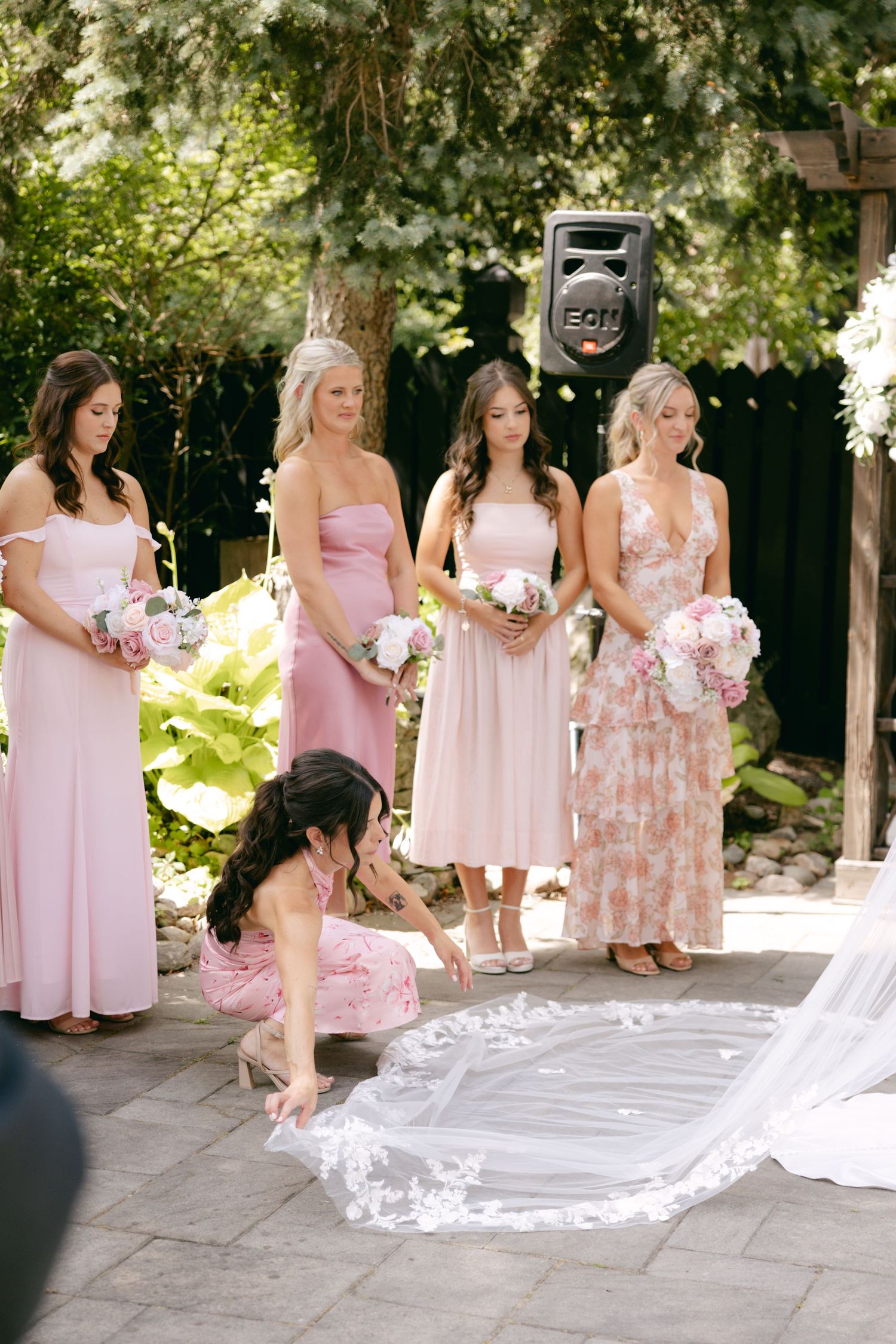 Bridesmaids in pink dresses arrange the bride's long veil on stone patio.