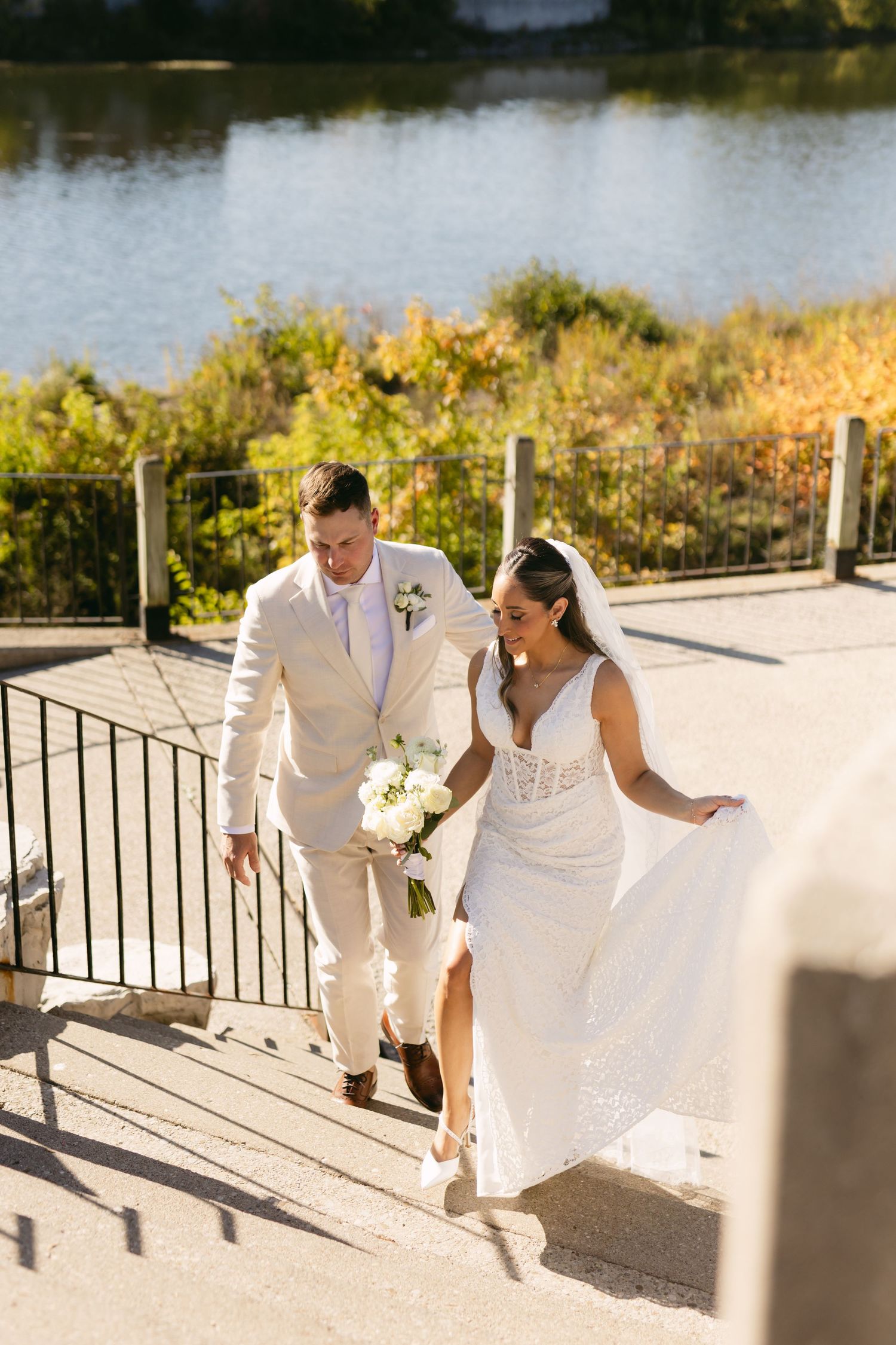 A couple in cream-colored wedding attire walks along a waterfront path during their photoshoot.