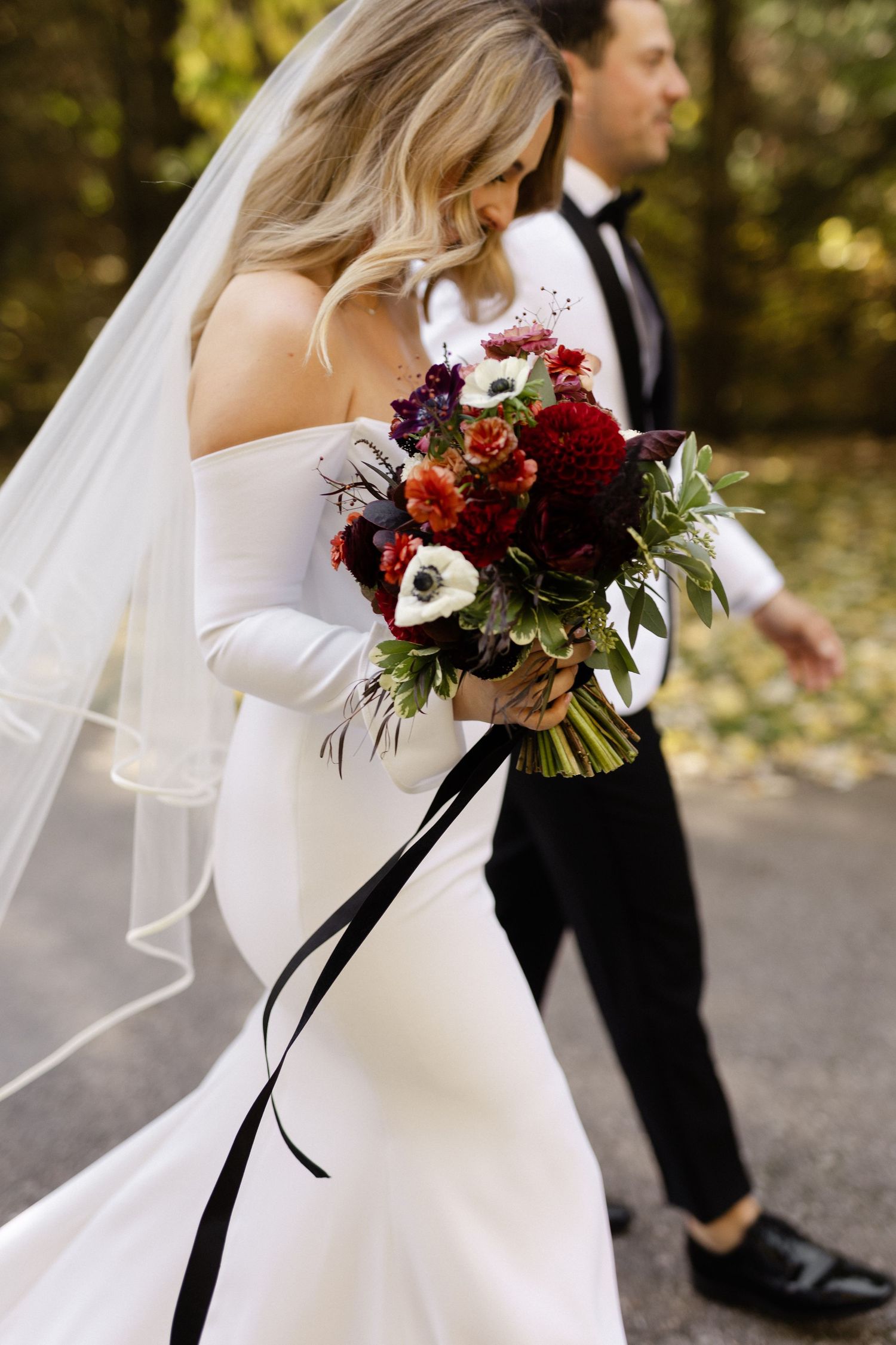 Bride in off-shoulder gown holds deep red bouquet with black ribbons.