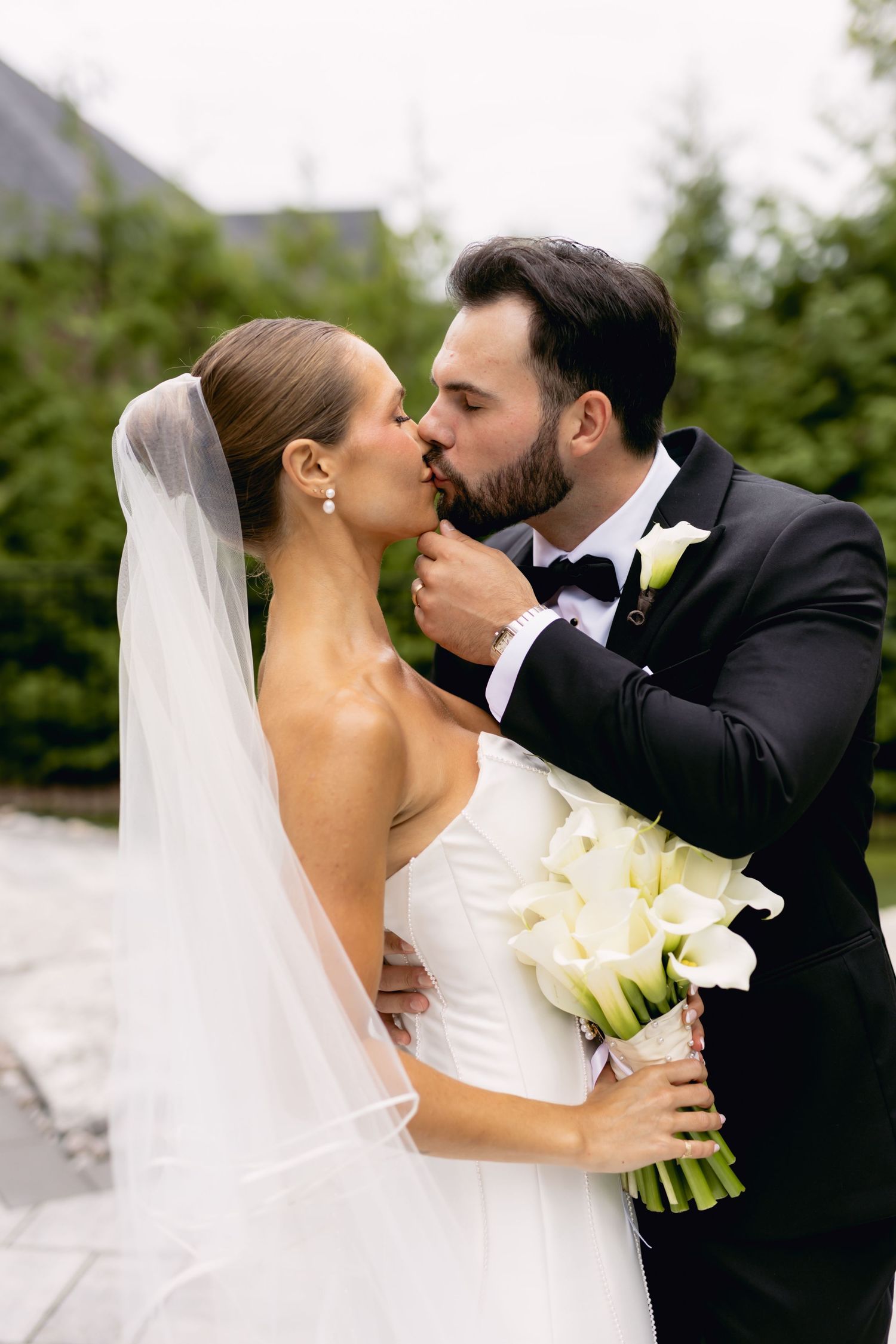 Groom in black tuxedo tenderly kisses bride in white strapless gown holding white calla lilies in garden setting.