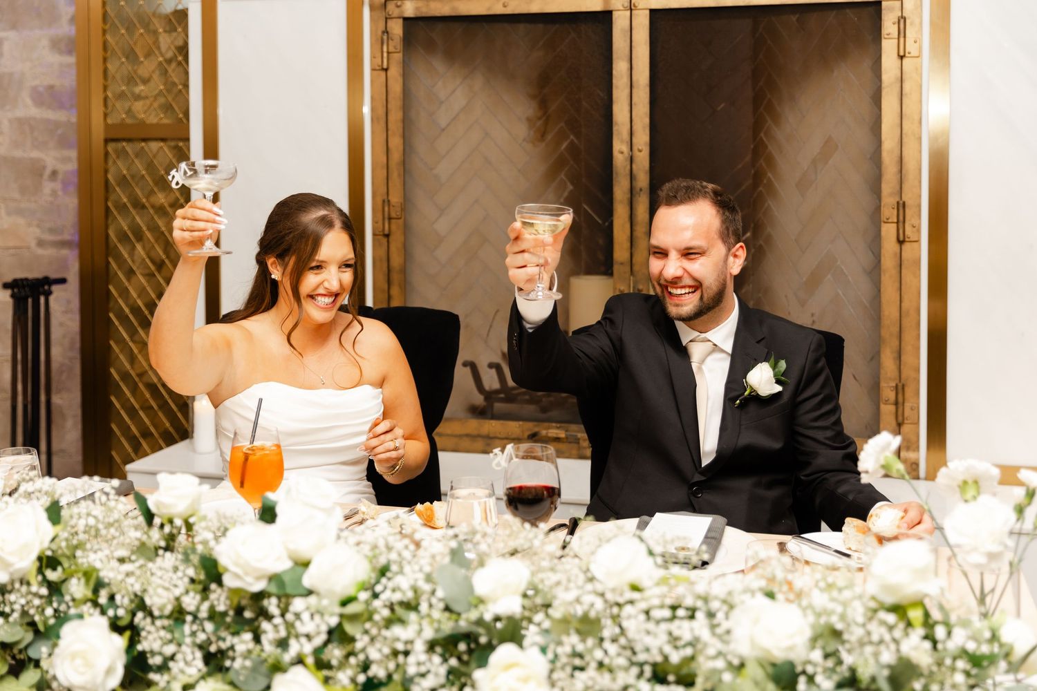 Smiling bride and groom raise glasses in toast at sweetheart table decorated with white florals.