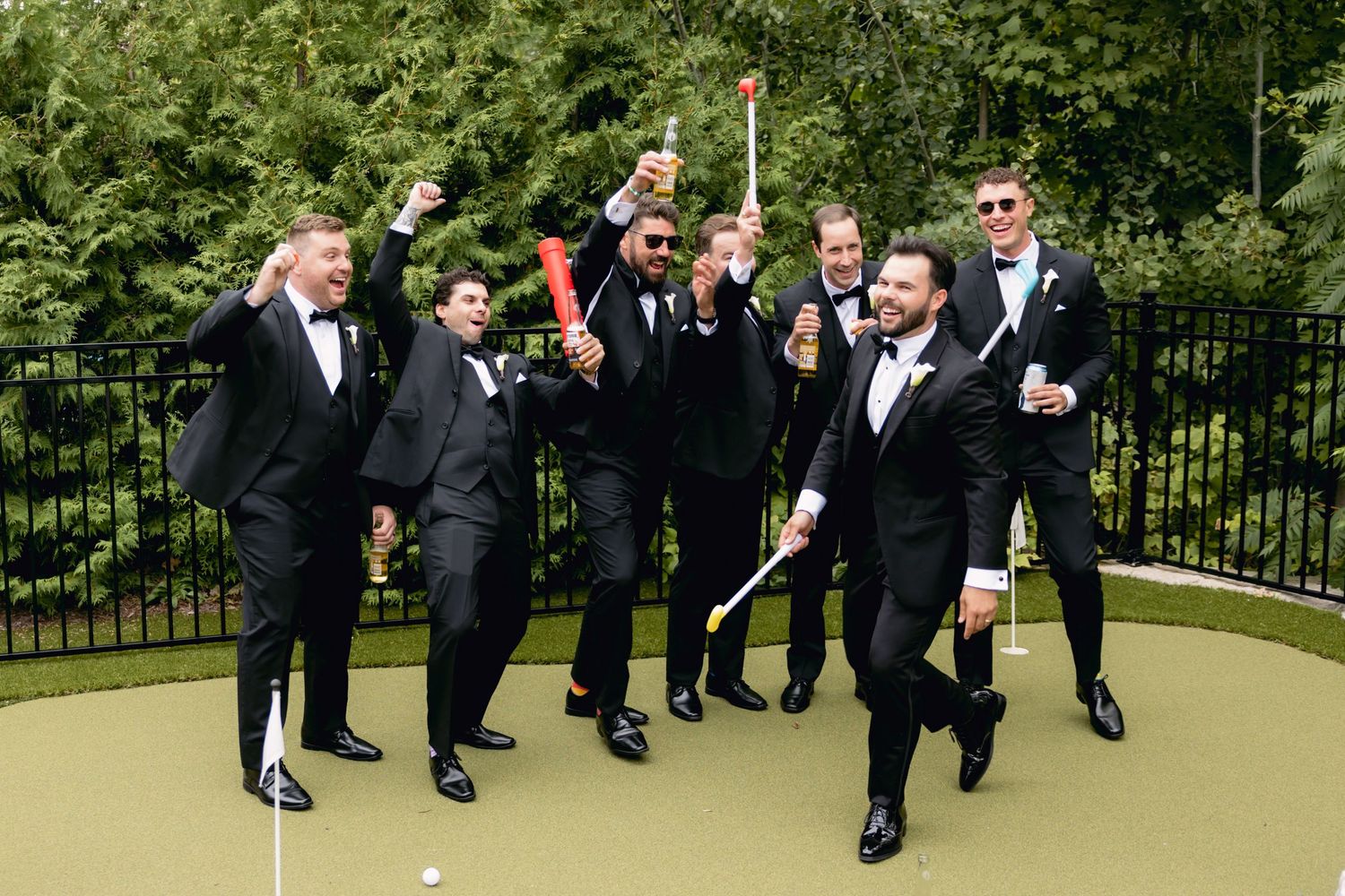 Energetic groomsmen in black tuxedos celebrate on a putting green, raising drinks and golf accessories outdoors.