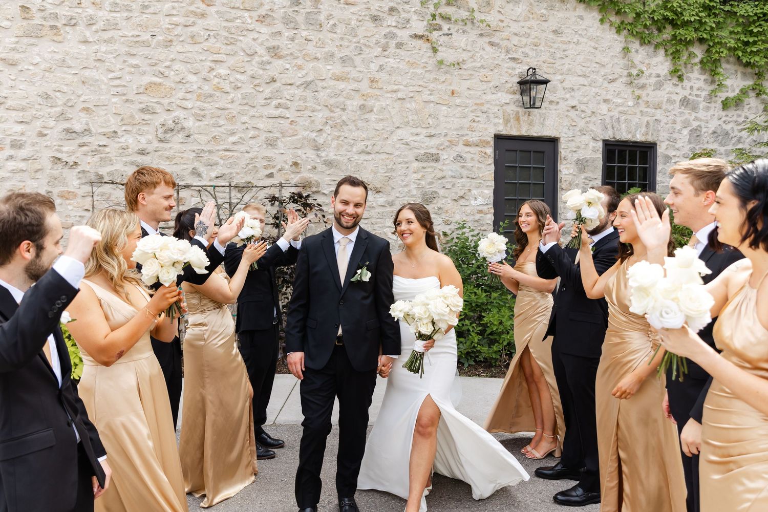 Bride and groom walk through cheering wedding party in gold bridesmaid dresses outside stone venue.