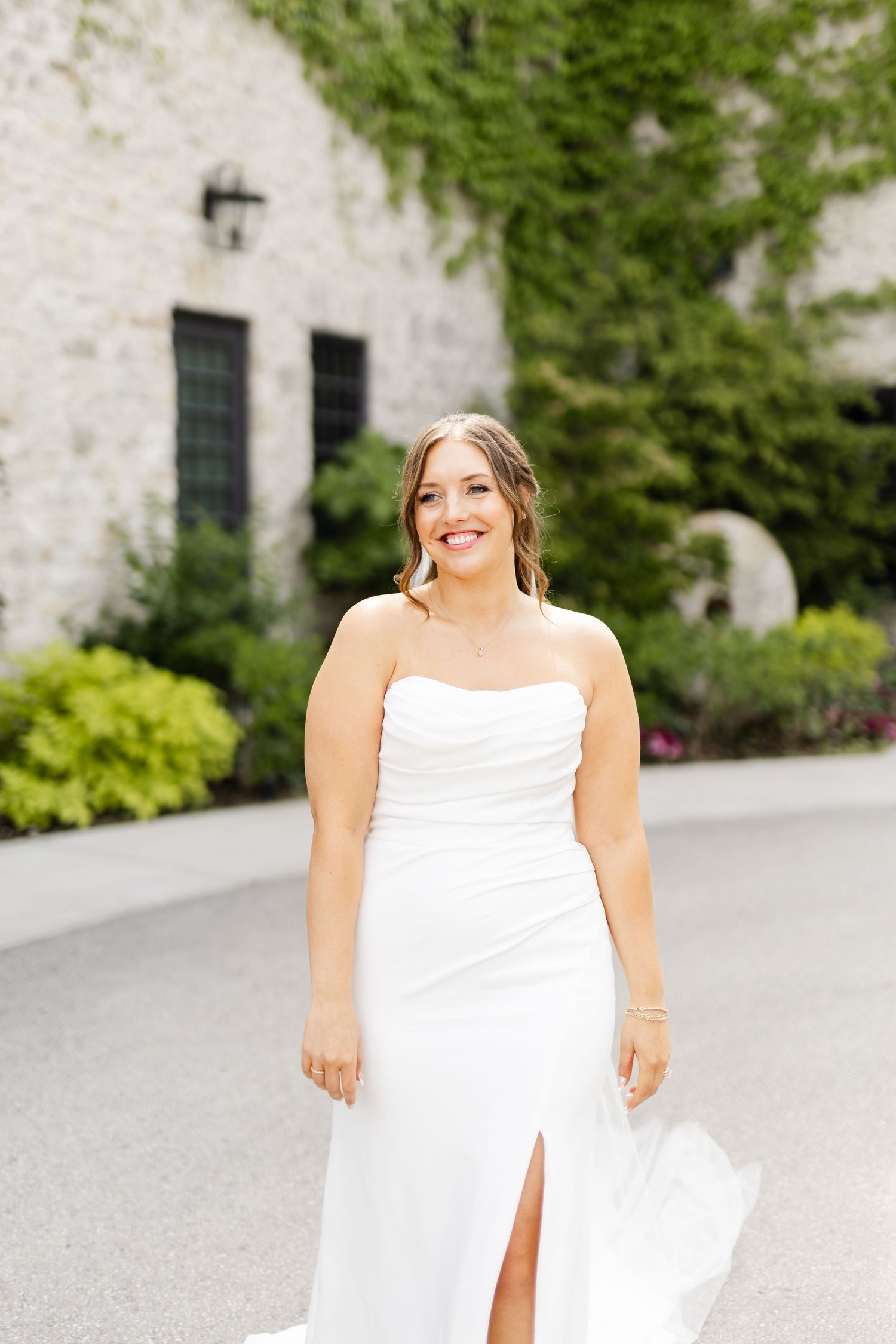 Smiling bride in a strapless white wedding gown with a front slit poses outdoors at a stone venue.