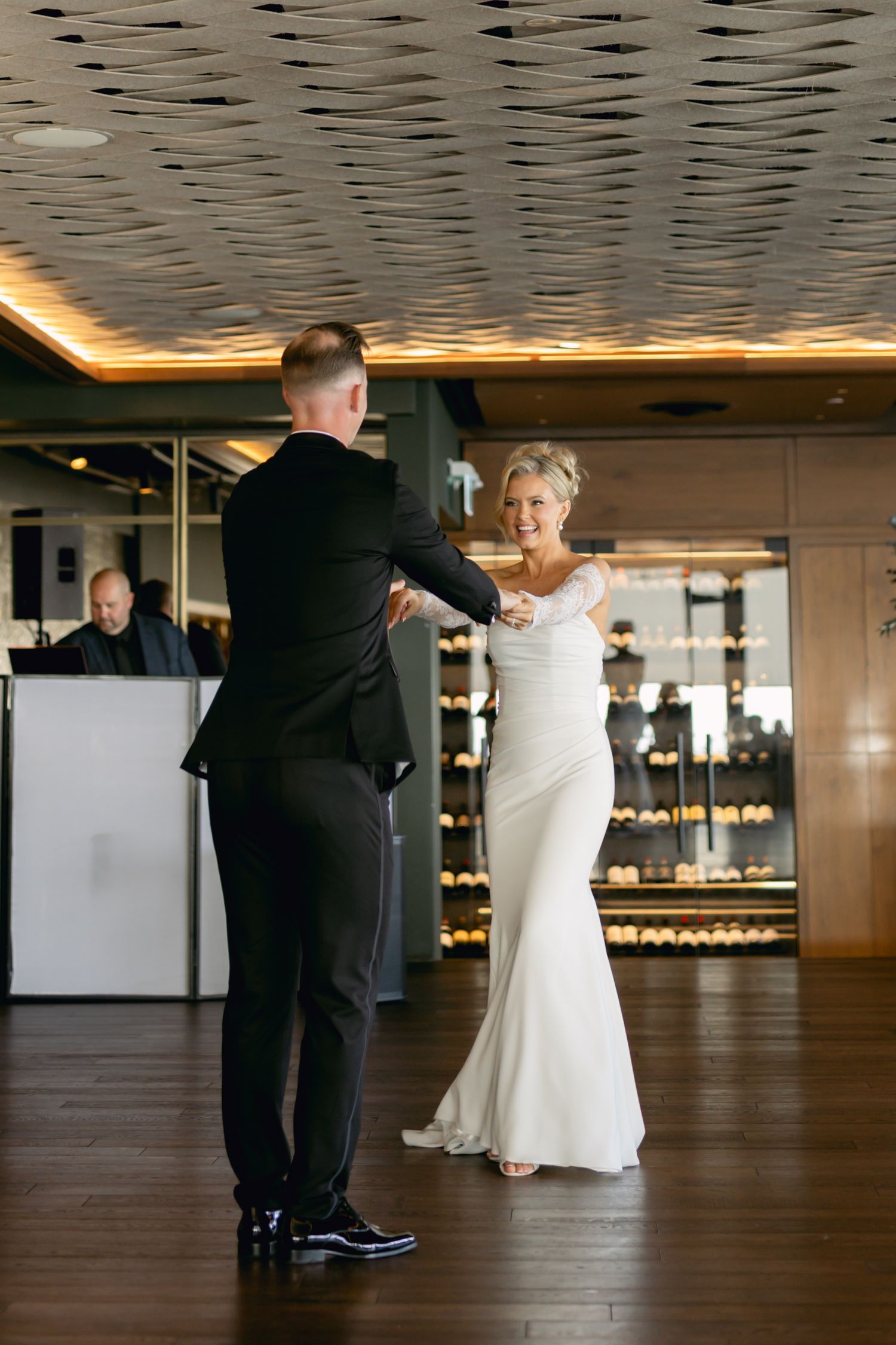 Bride in fitted white lace gown and groom in black tuxedo sharing first dance on hardwood floor.