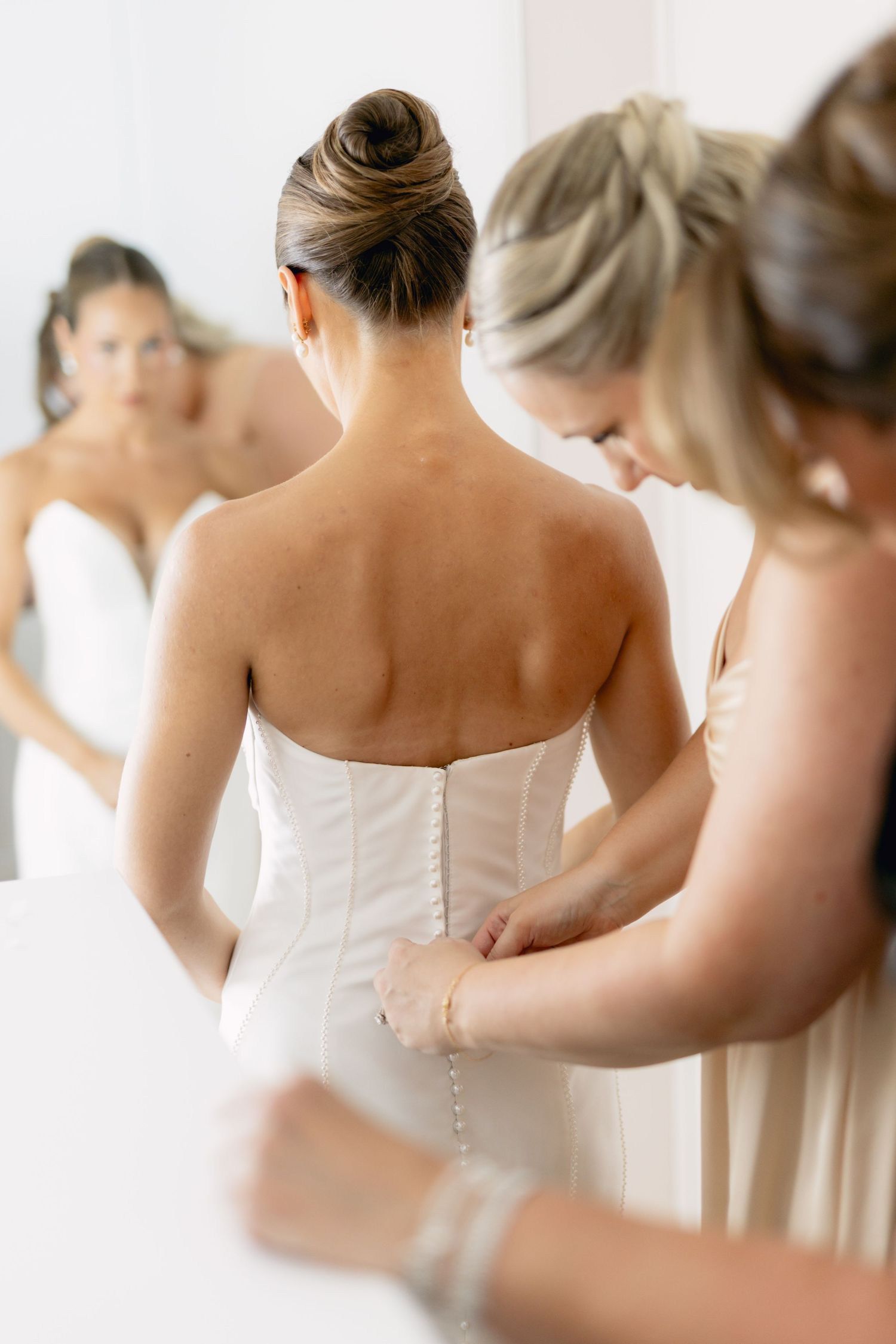 Bridesmaid buttoning the back of a strapless white wedding gown while reflected in a mirror.