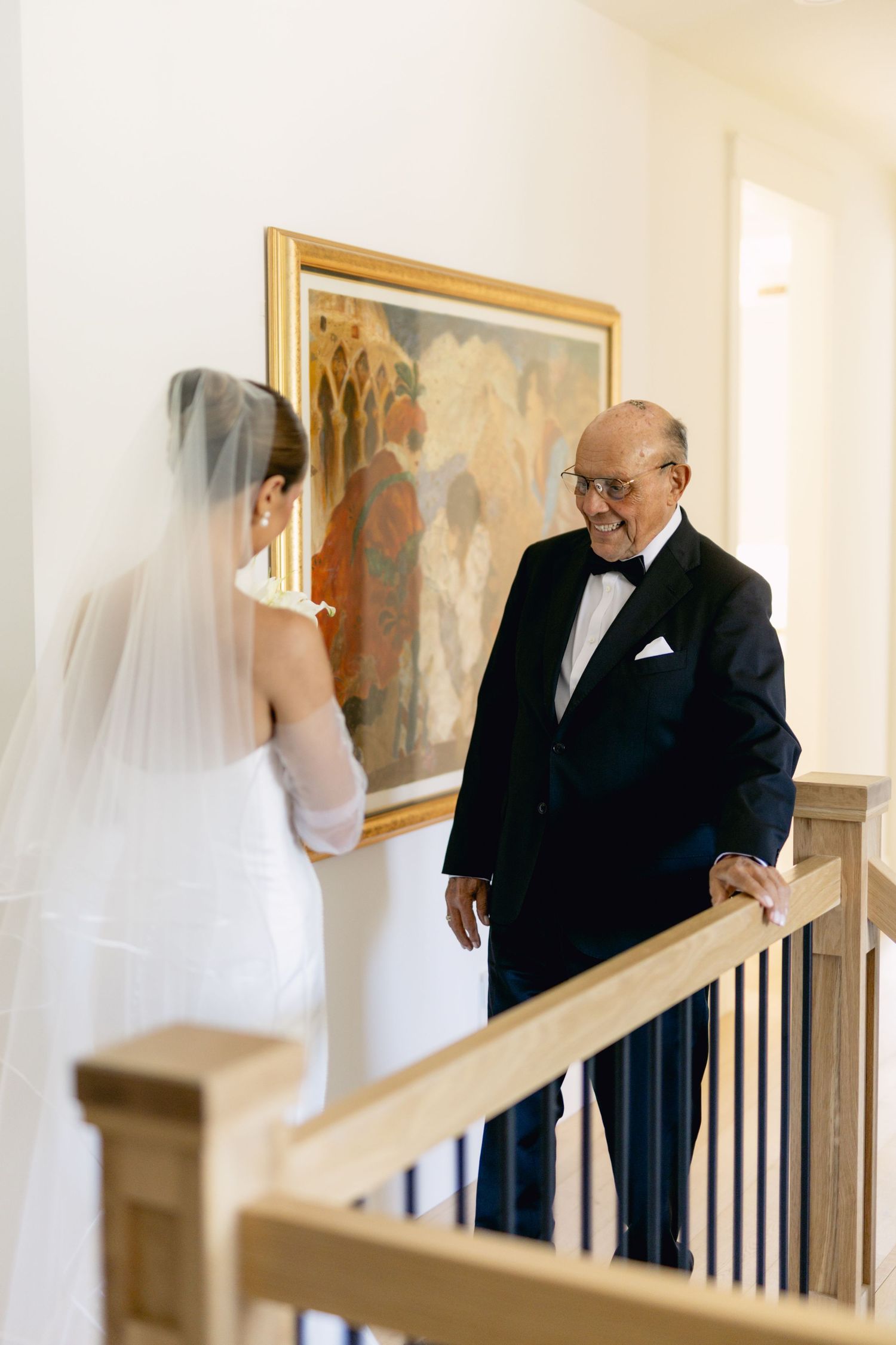 Emotional father in tuxedo seeing bride in white veil and gown for first time on staircase.