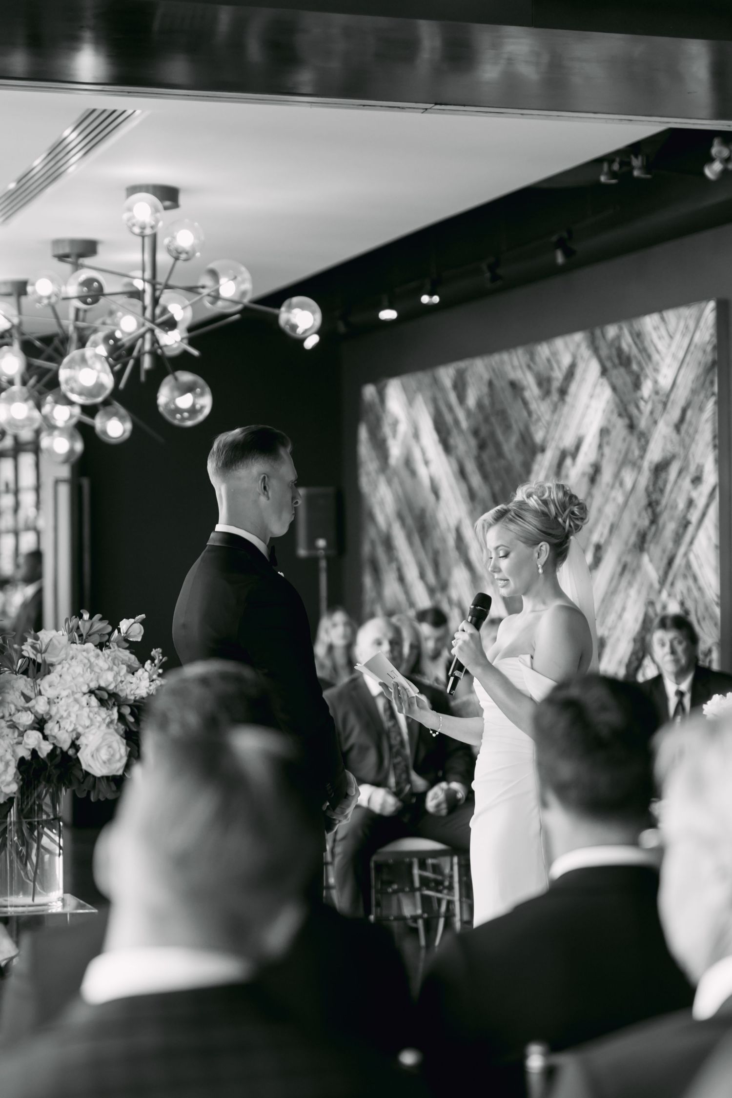 Black and white photo of bride reading vows to groom before seated wedding guests under modern chandelier.