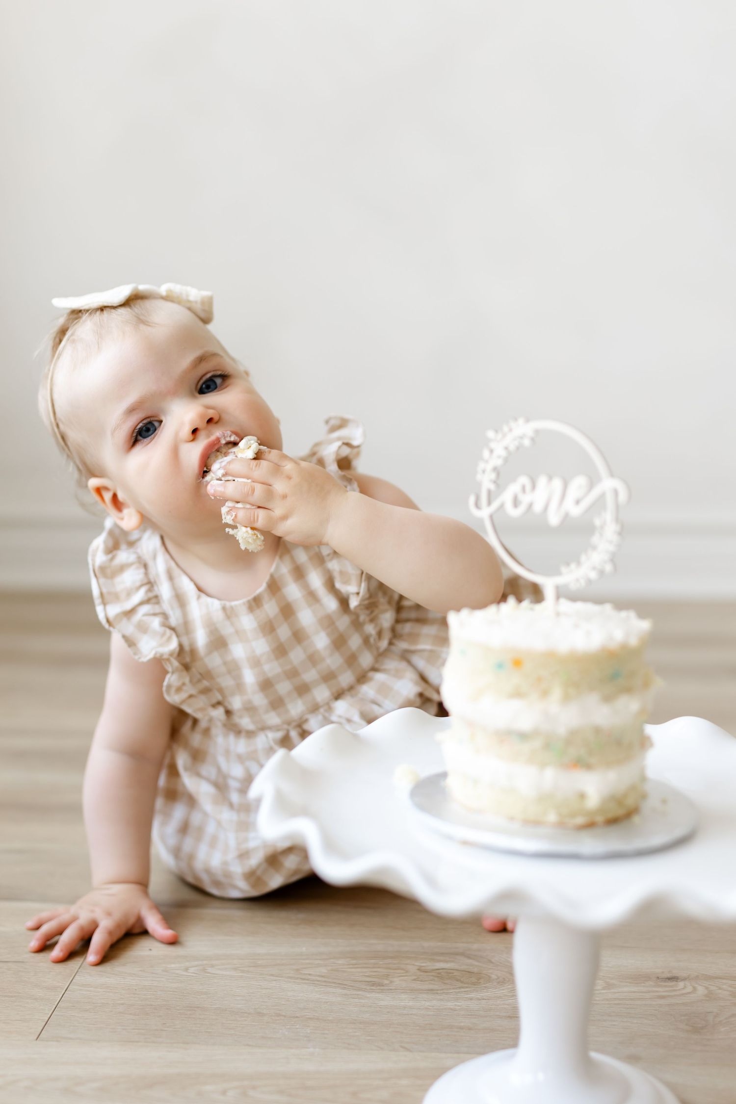 Baby girl in gingham dress smashing her first birthday cake, with a white 'one' cake topper on a pedestal stand.