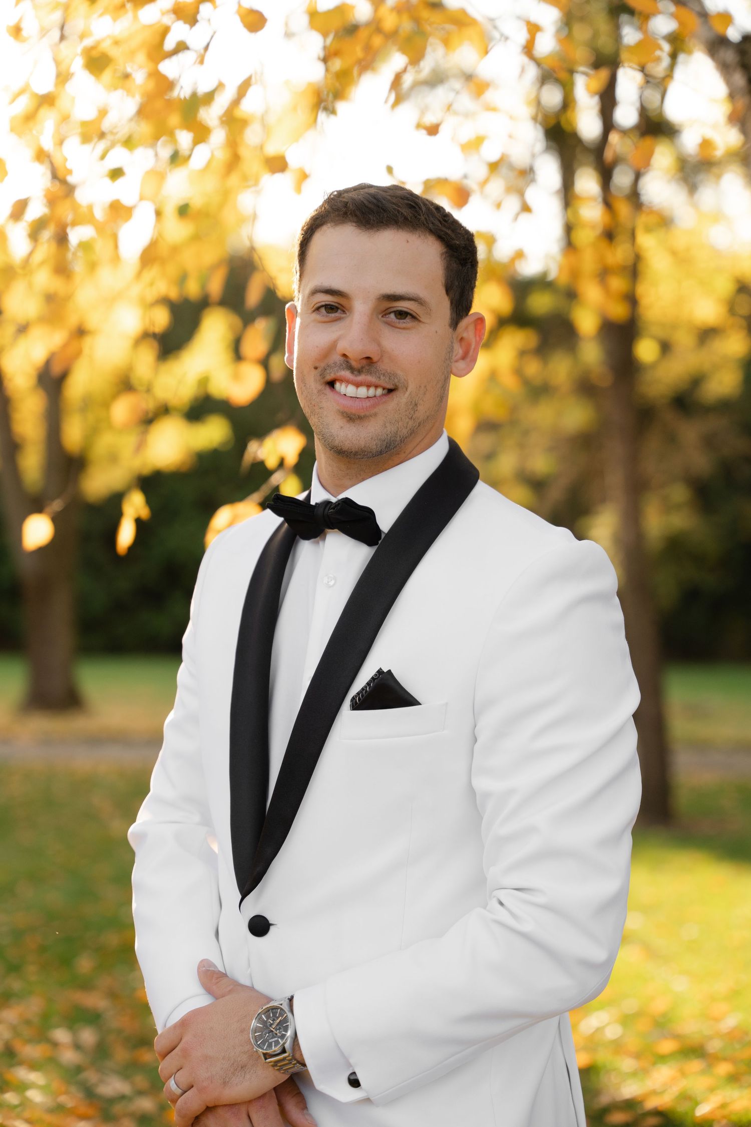 Groom in white tuxedo jacket and black bow tie poses outdoors during golden hour autumn wedding photoshoot.