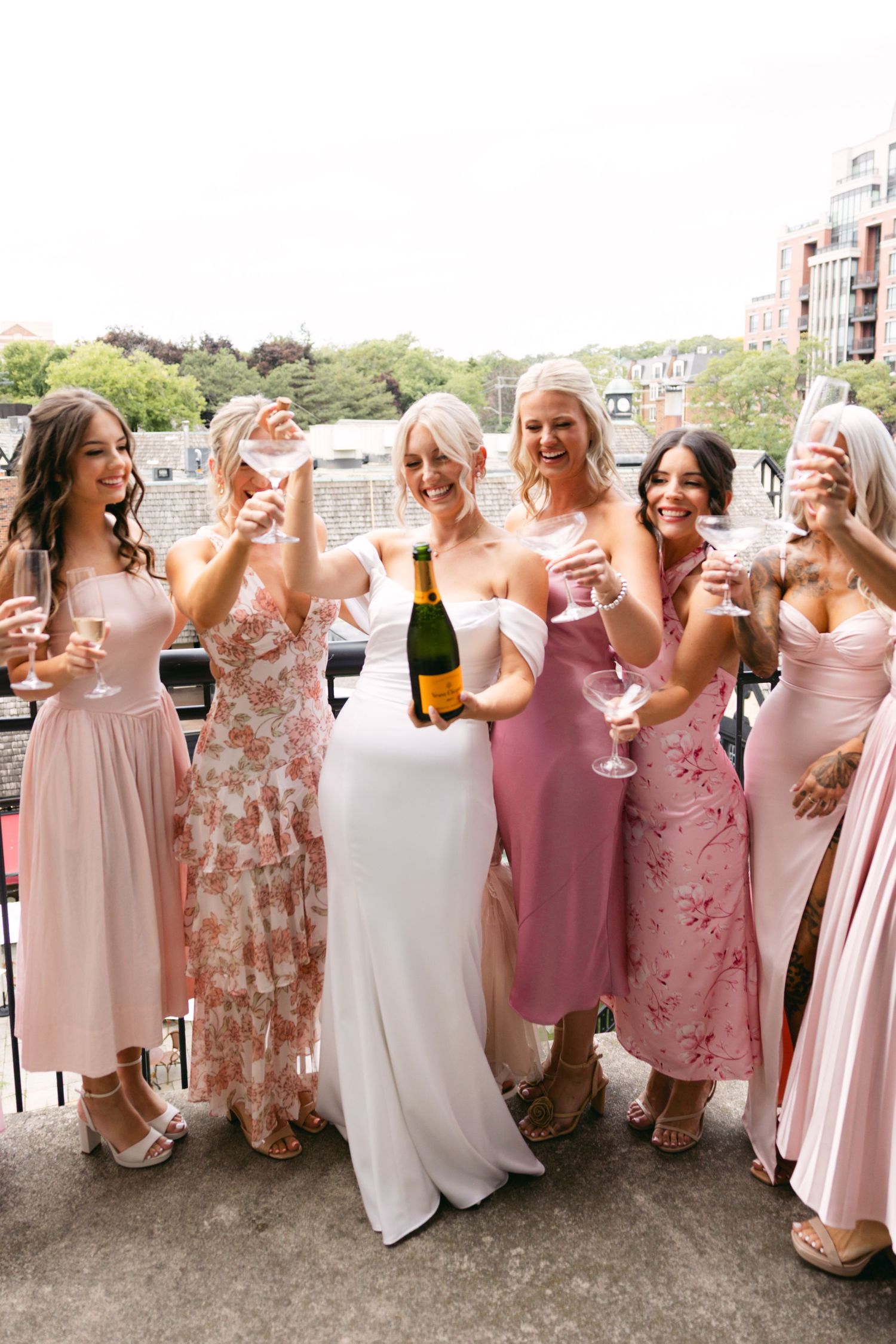 Bridal party in pink and floral dresses celebrating with champagne on outdoor terrace.