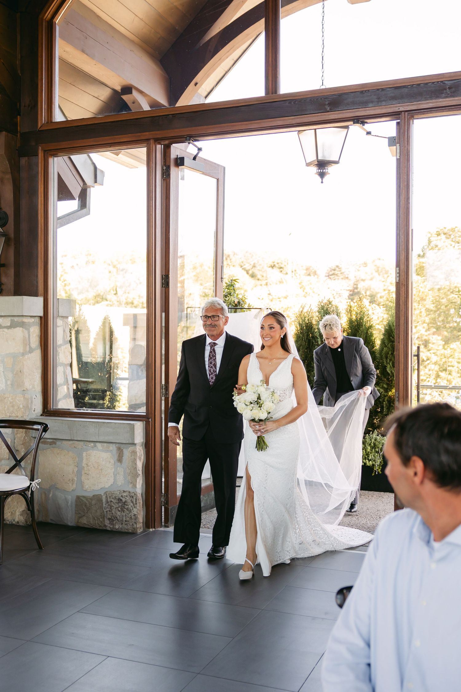 A bride in a flowing white gown enters a rustic wedding venue with large windows and stone walls.