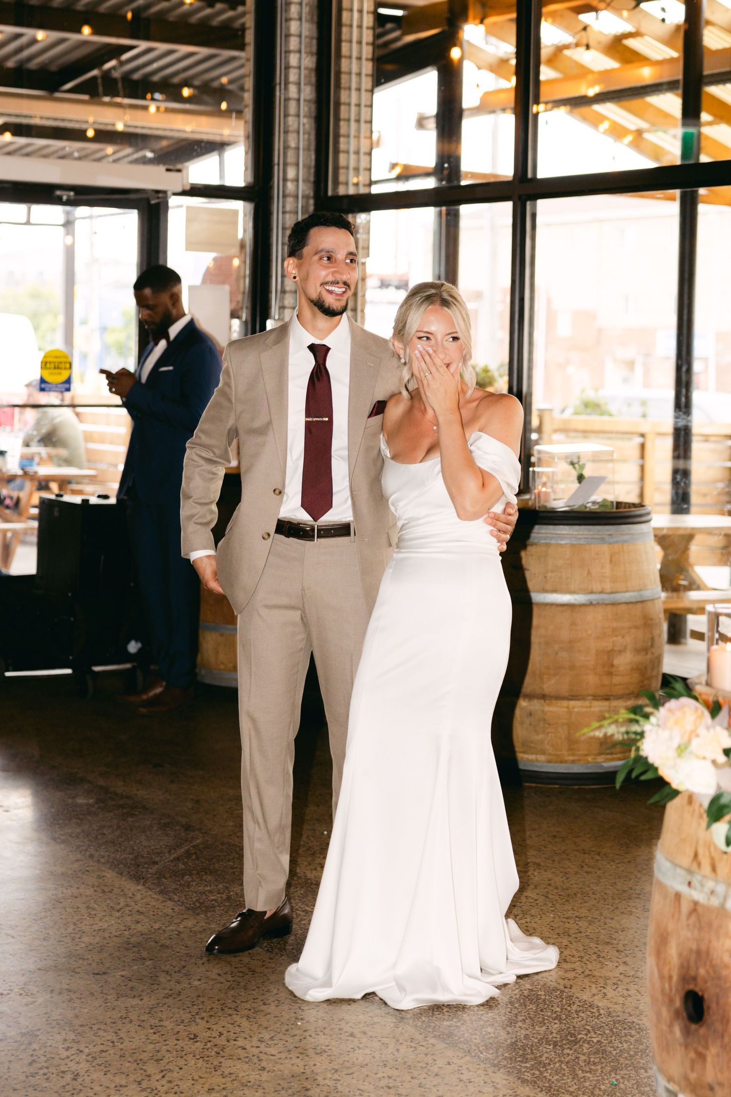 Wedding party celebrates in rustic venue with wooden barrels and natural lighting.