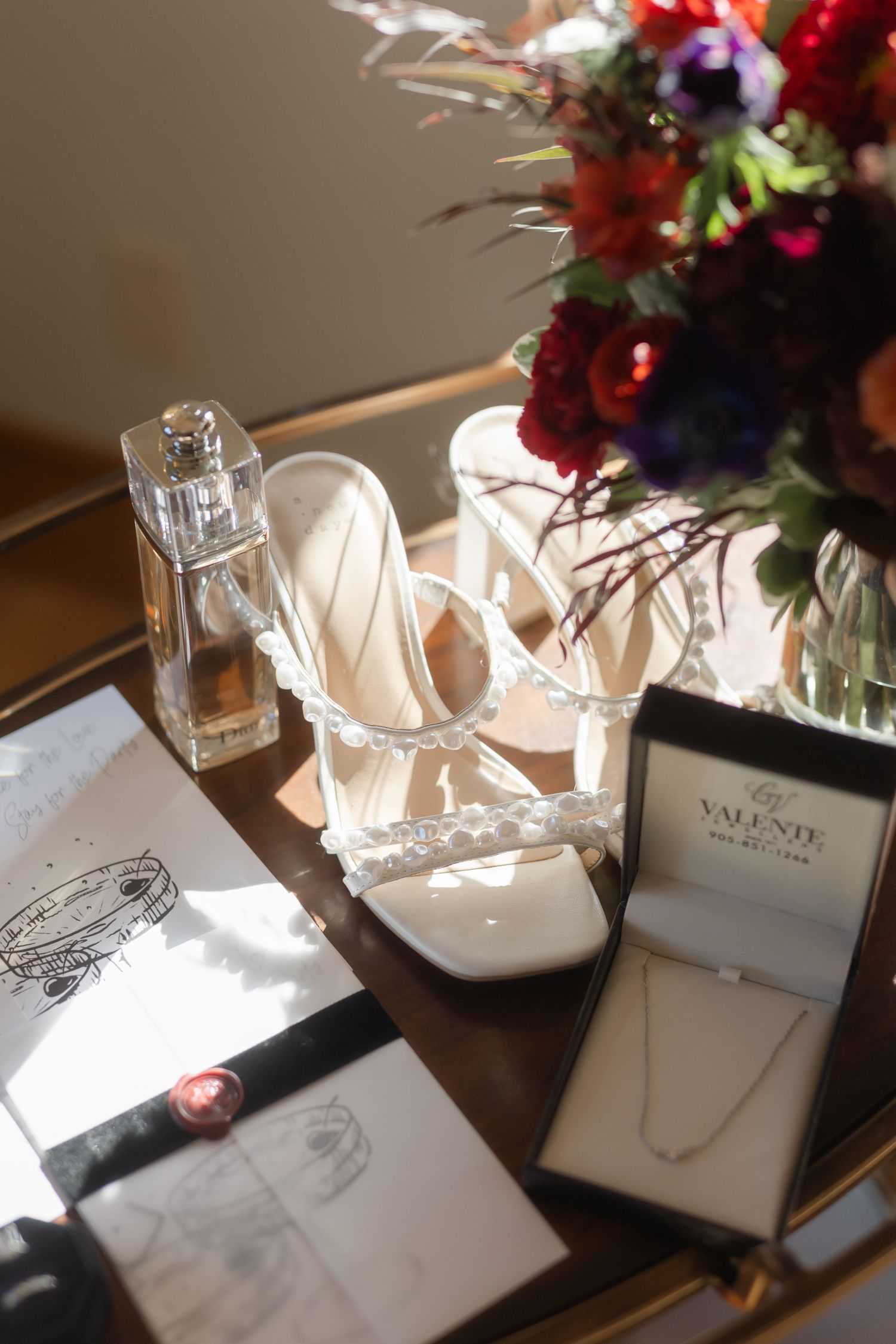 Wedding accessories including white heels, jewelry box and vibrant red floral arrangement on wooden surface.